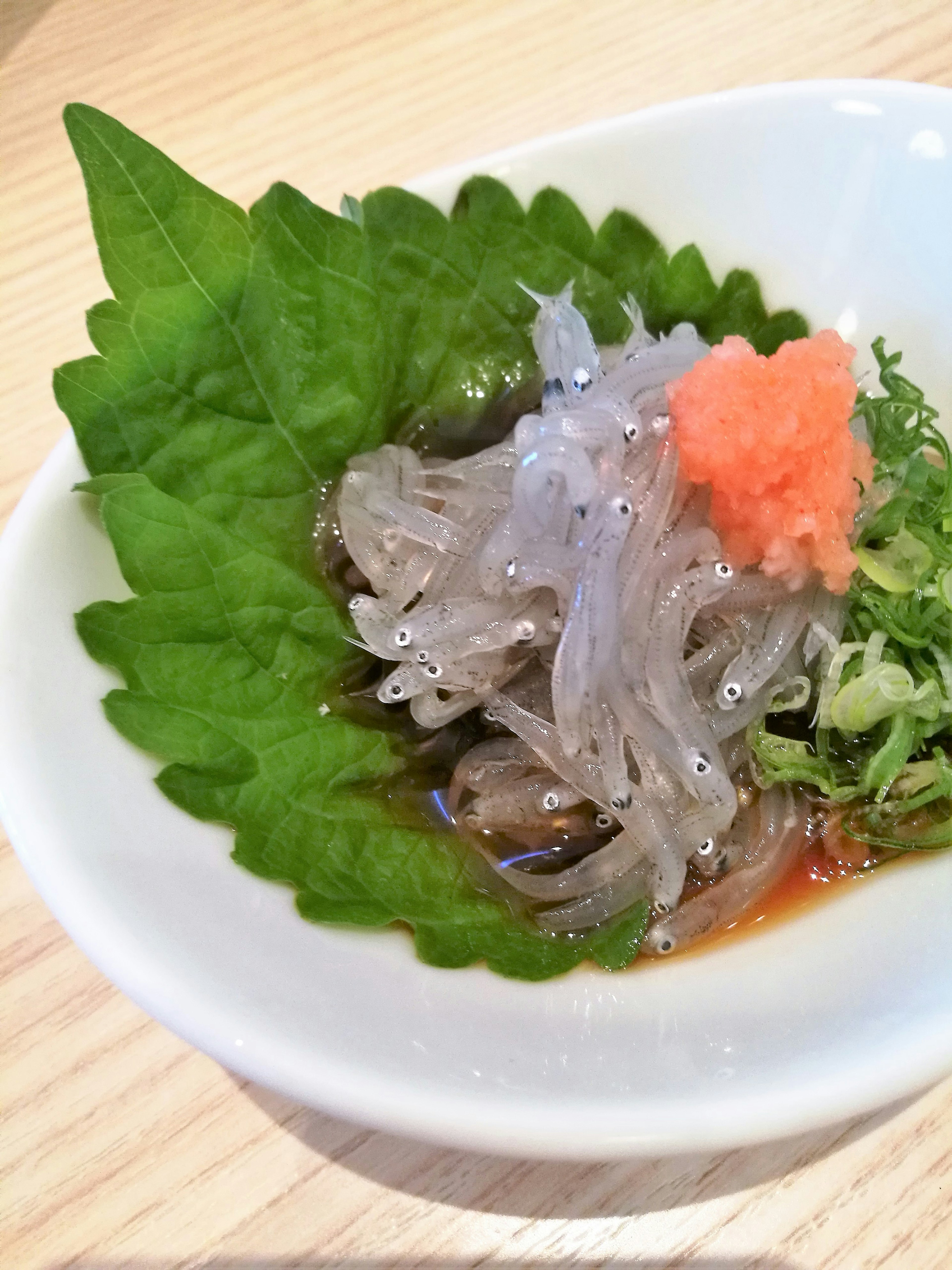 A small dish featuring translucent shirasu sashimi garnished with perilla leaves, green onions, and pickled ginger