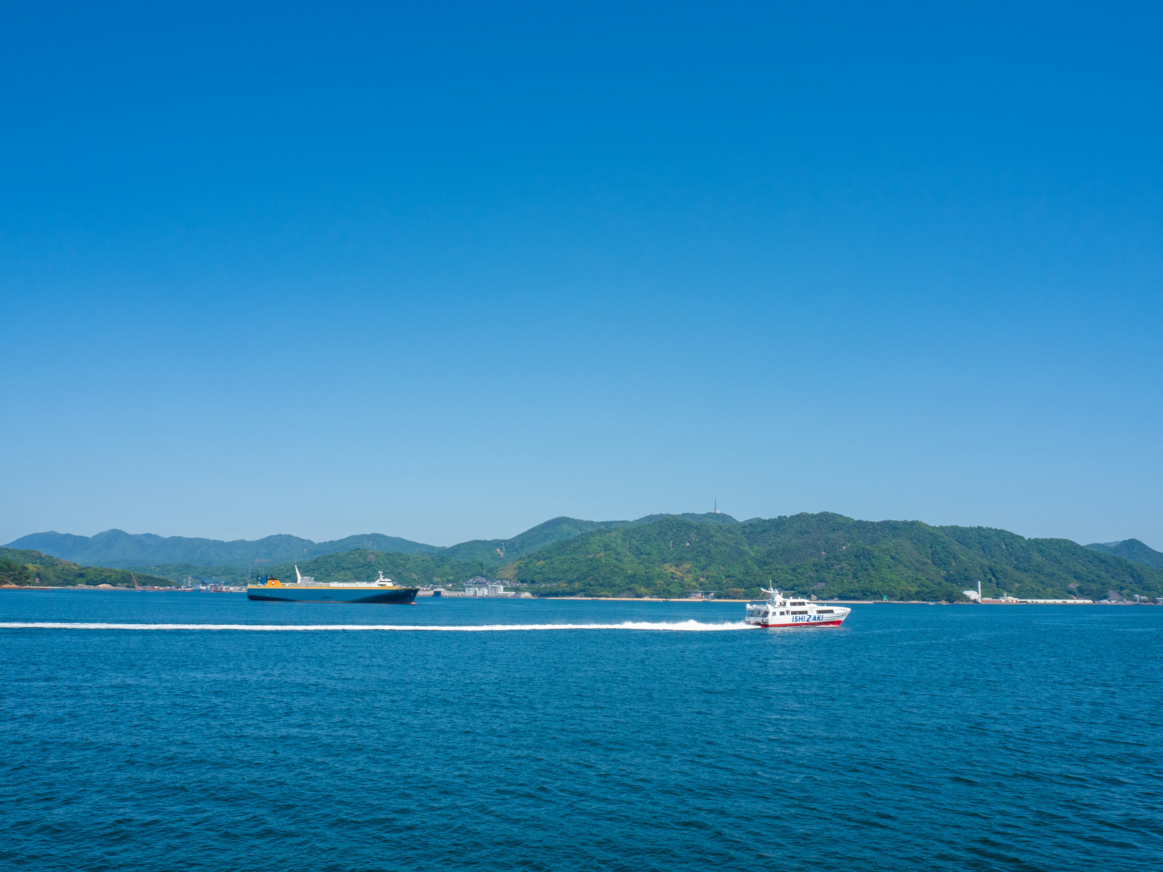 Eine malerische Aussicht auf ein blaues Meer mit einem kleinen Boot und einem großen Frachtschiff vor einer bergigen Kulisse