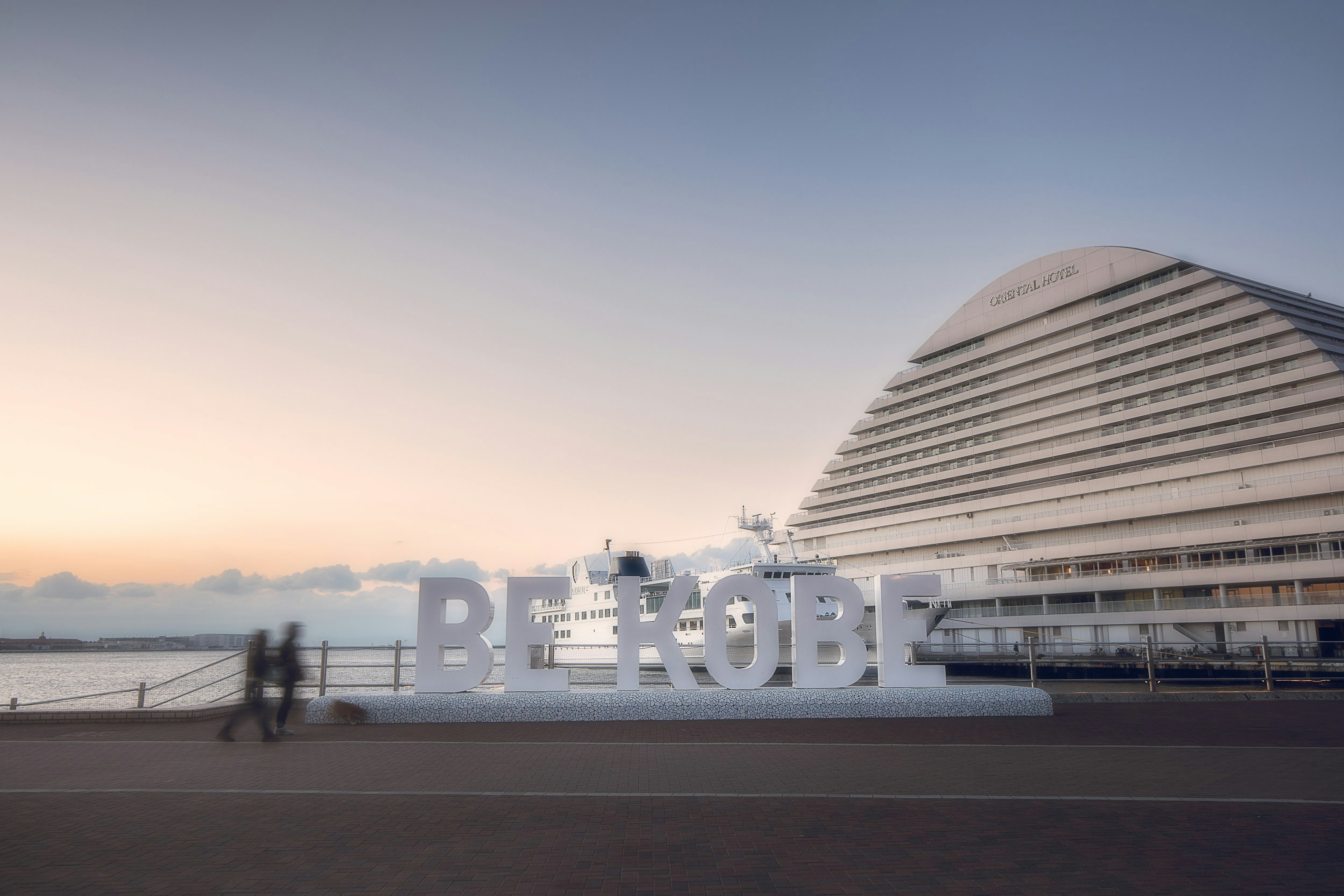 Grandes letras blancas que dicen 'BE RODE' cerca del mar con un edificio moderno al fondo