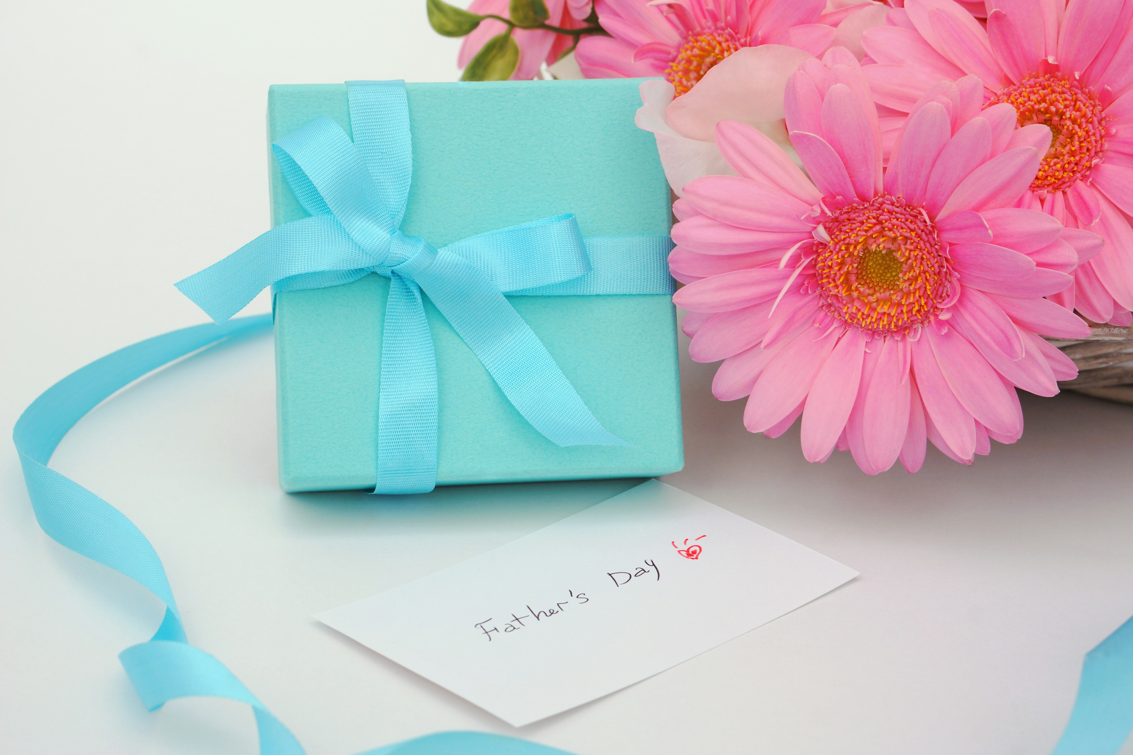 A blue gift box with a ribbon beside a bouquet of pink flowers and a greeting card