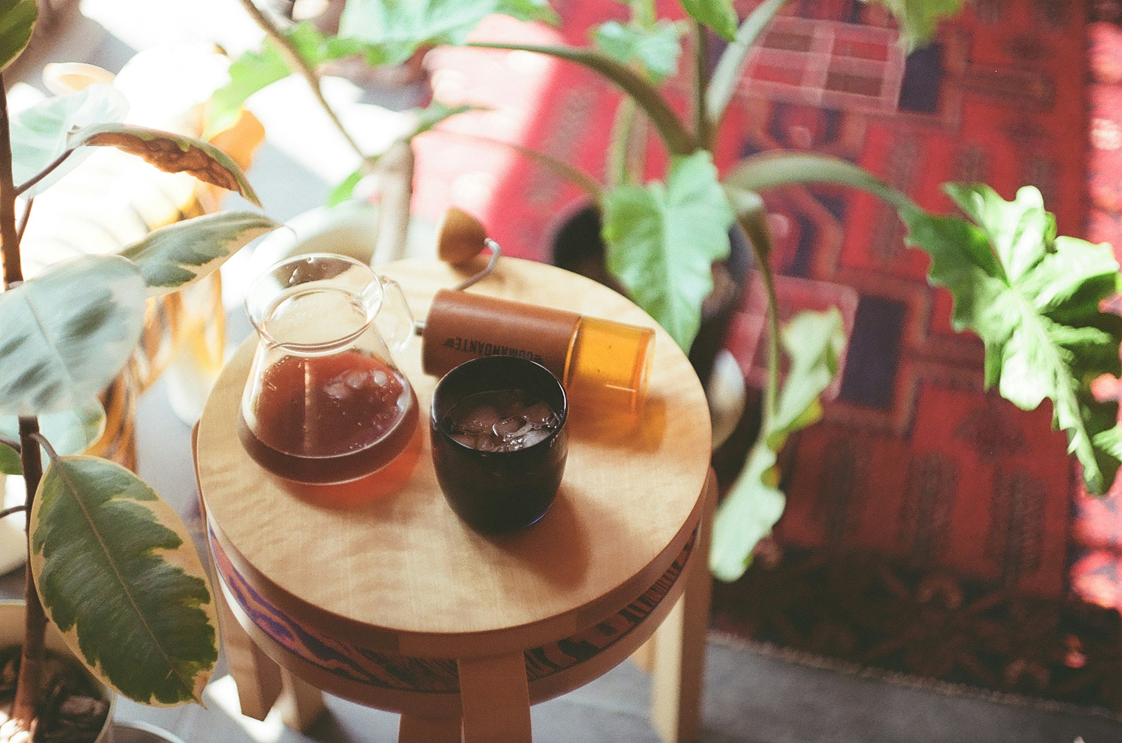 木製のスツールの上に置かれた茶器と飲み物のセット 緑の植物が背景にあり カラフルなラグが見える