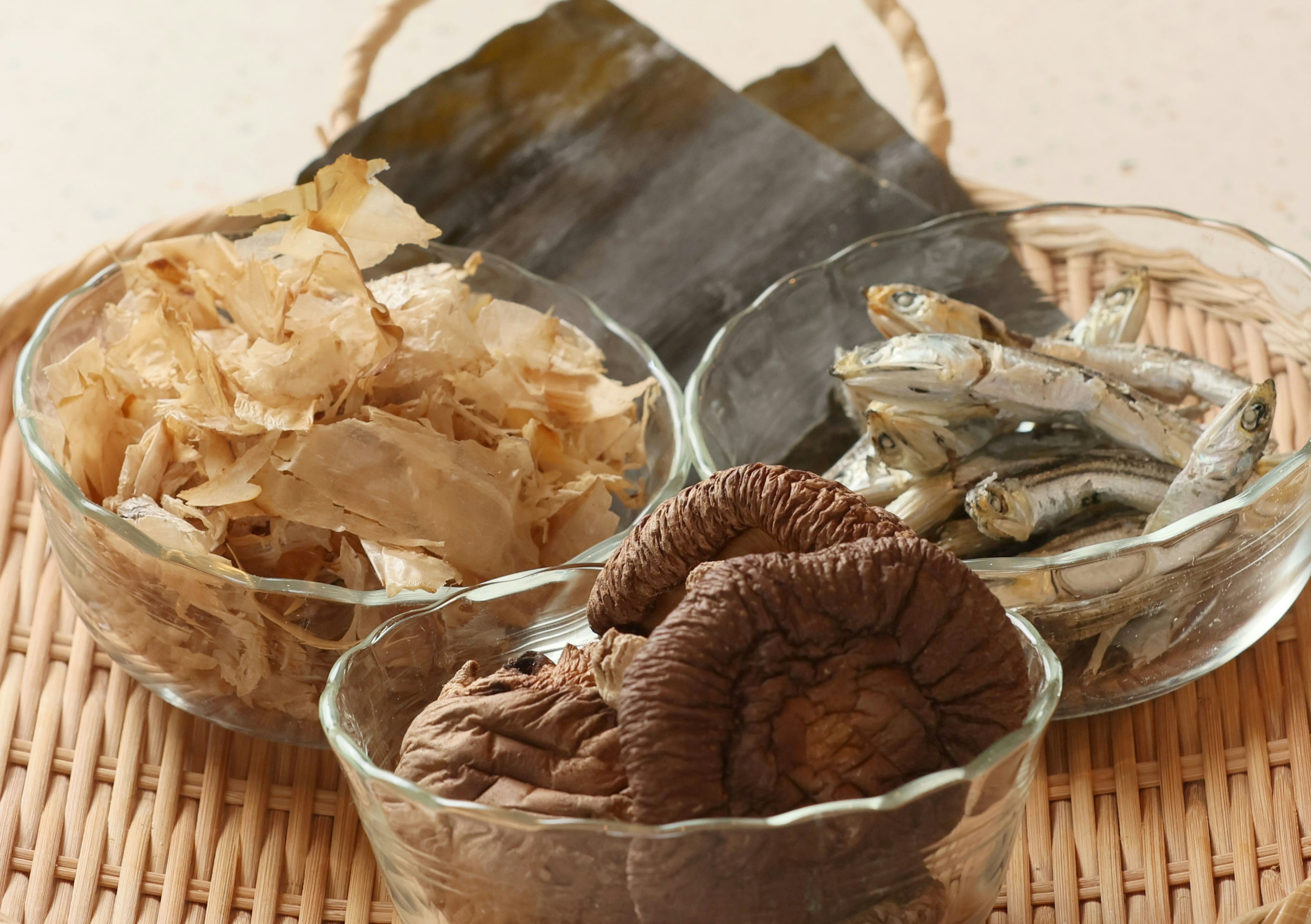 Bowls of dried bonito flakes, dried shiitake mushrooms, dried fish, and kelp on a woven mat