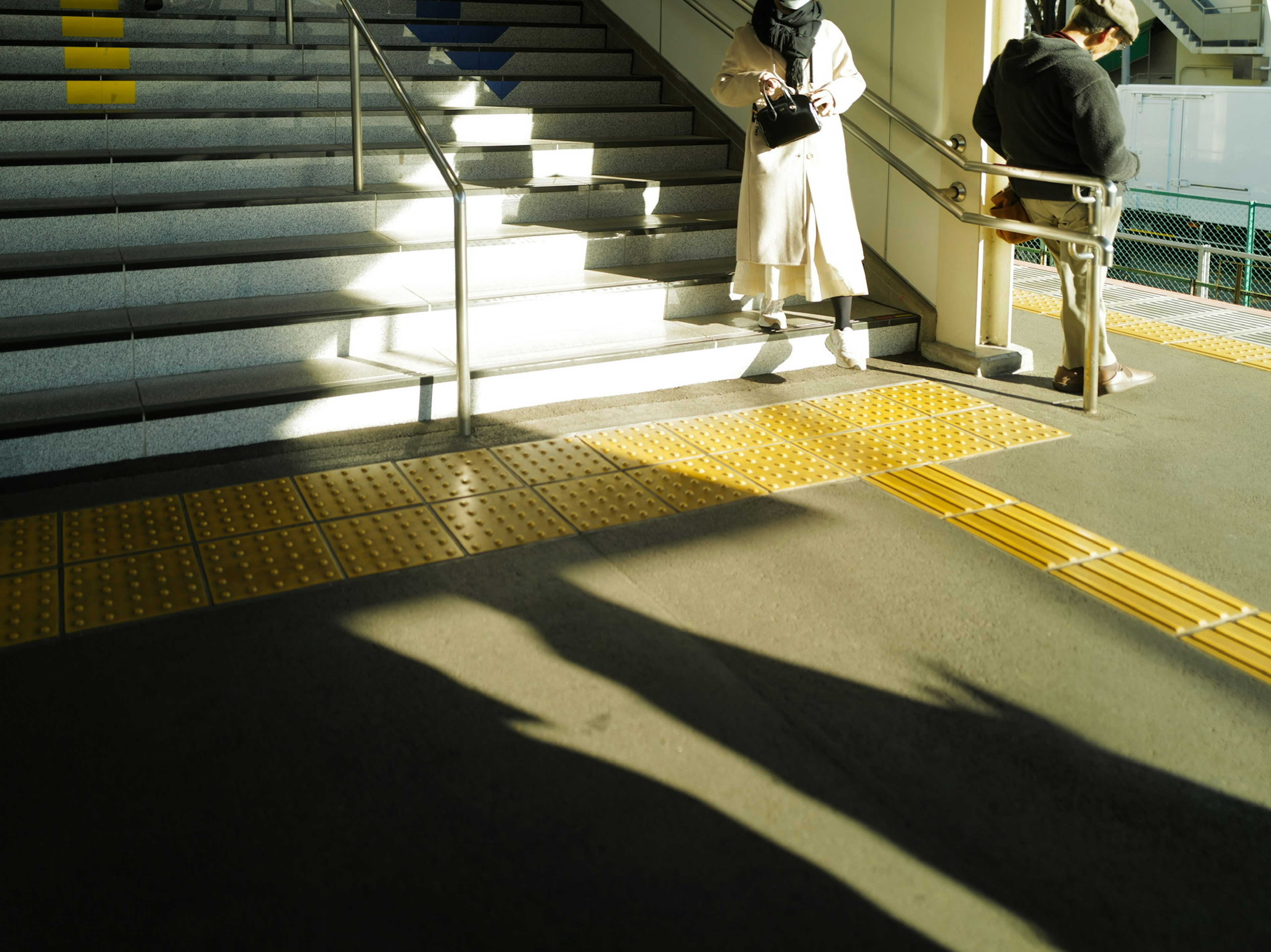 Eine Bahnhofszenen mit einer stehenden und einer sitzenden Person in der Nähe der Treppe, die Schatten werfen