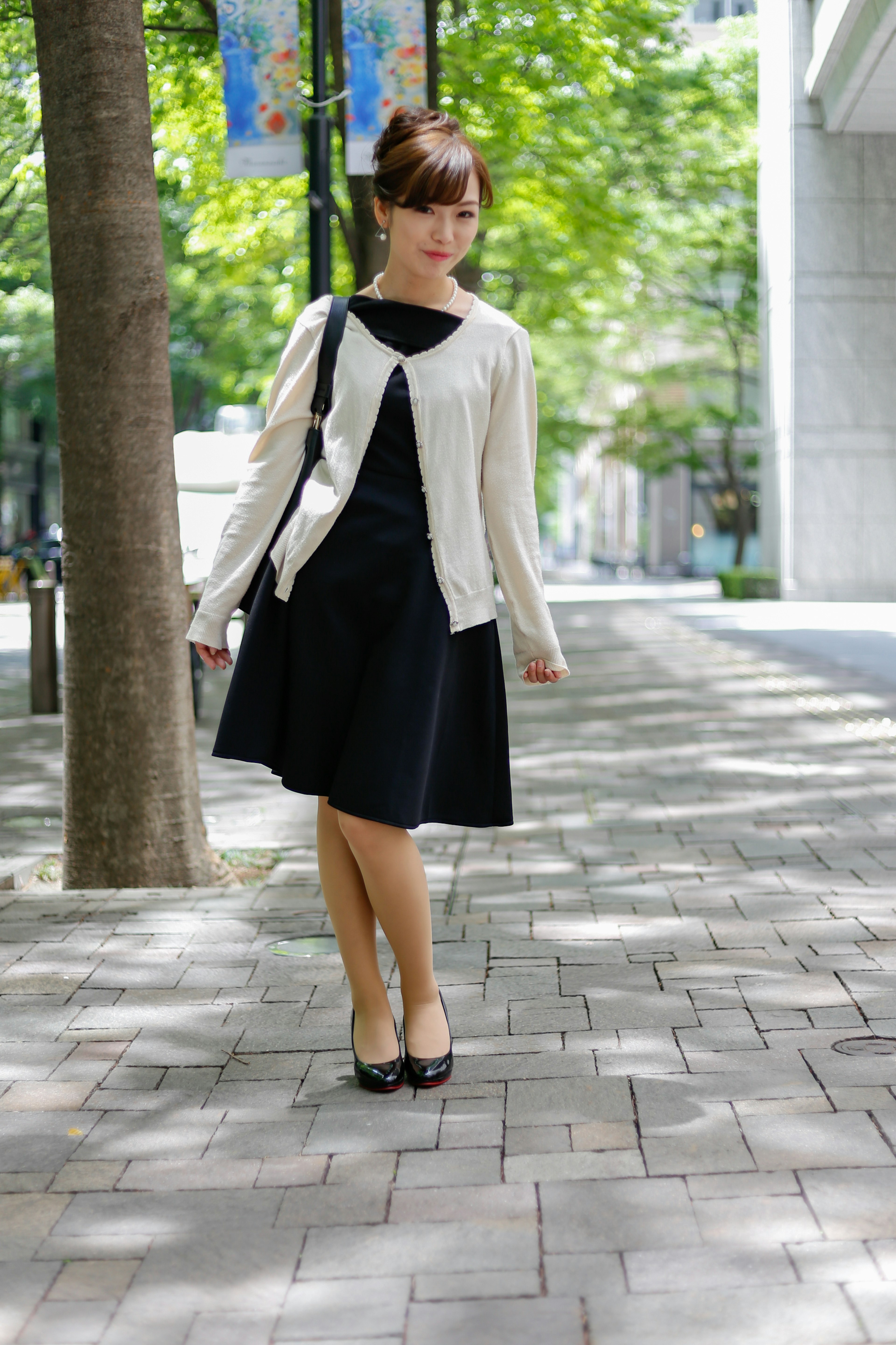 A woman stands on a city sidewalk wearing a white cardigan and black dress