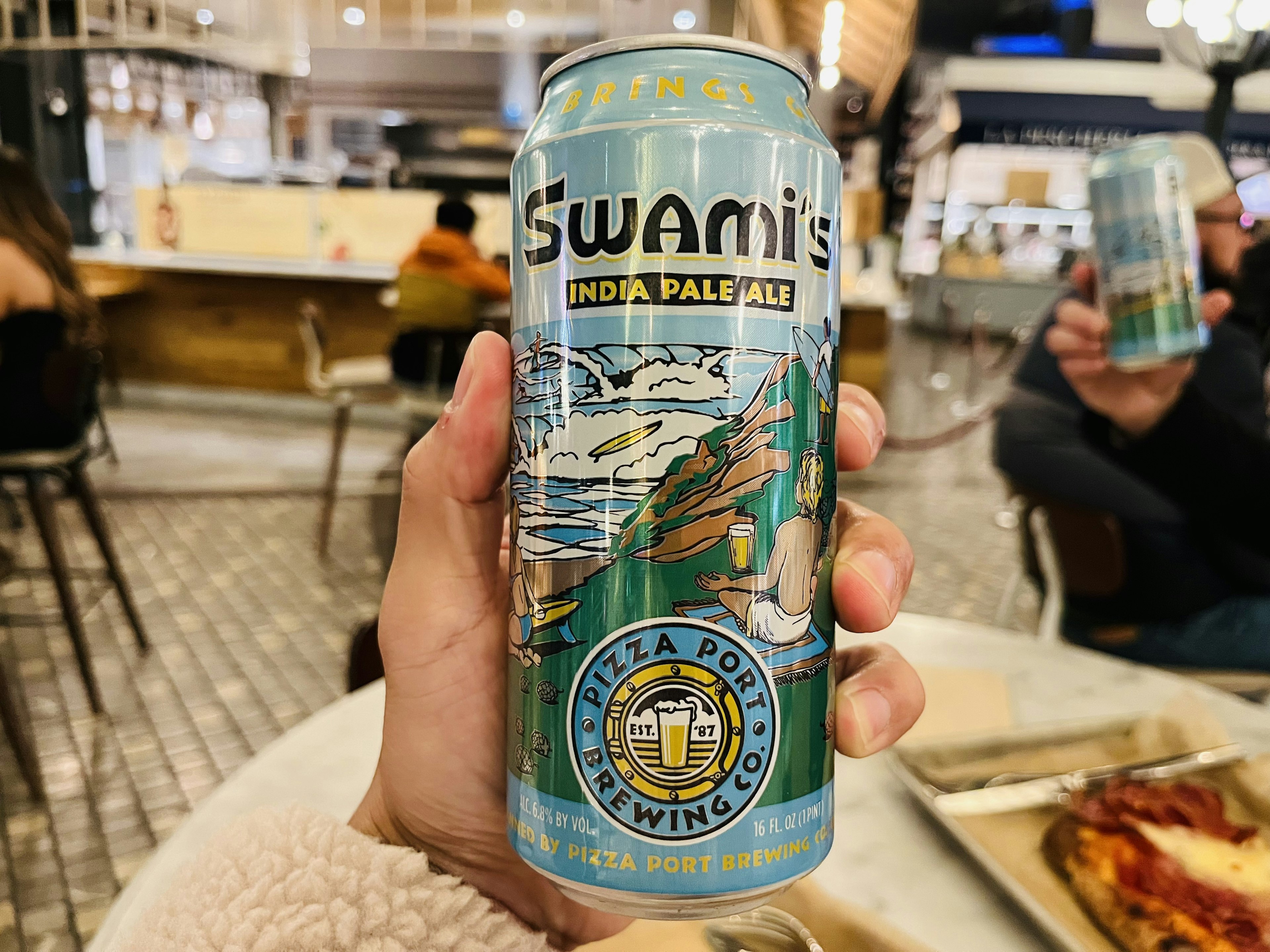 Hand holding a Swami's IPA can with a cafe background