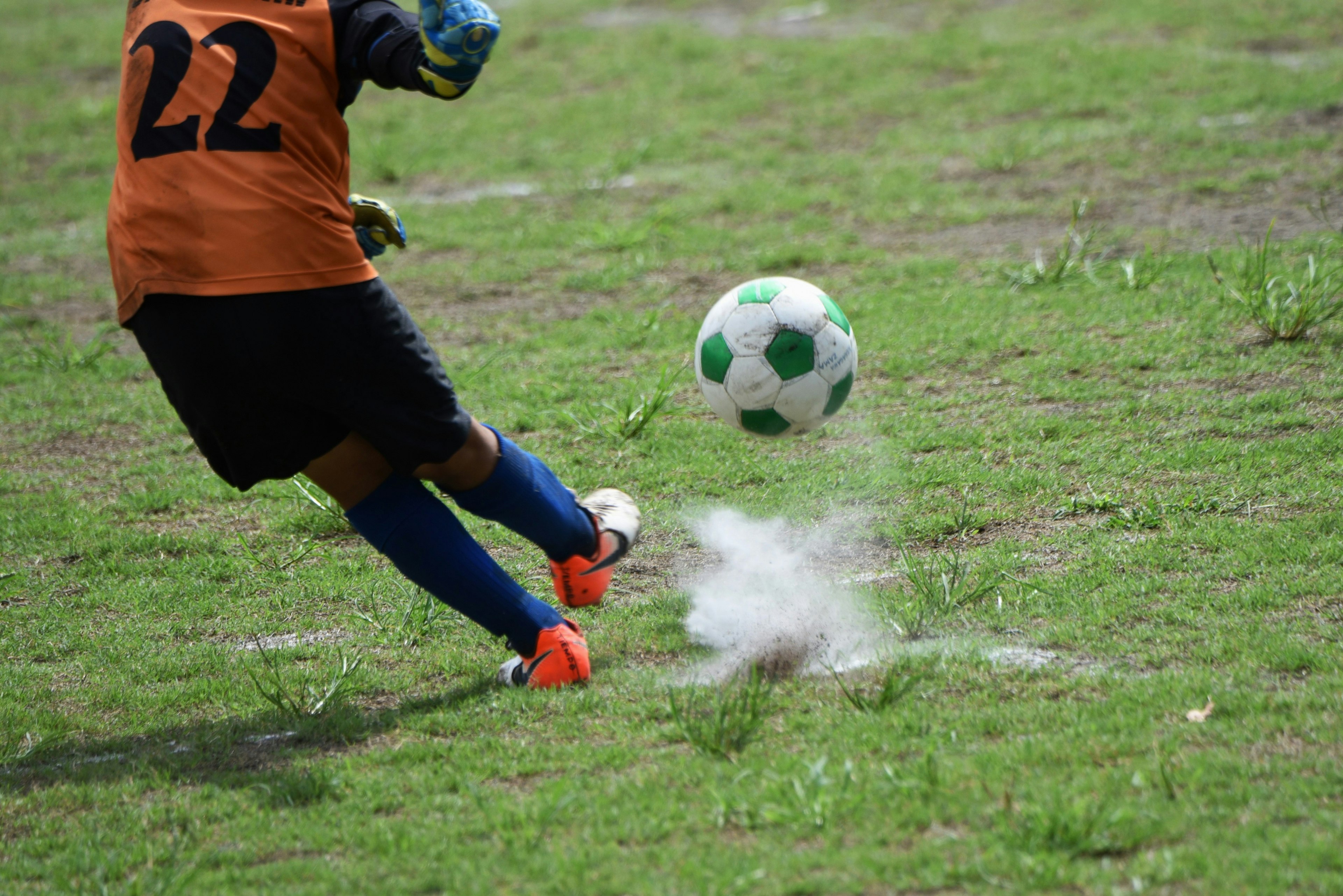 Jugador de fútbol pateando un balón en movimiento con una camiseta naranja con el número 22 sobre césped verde