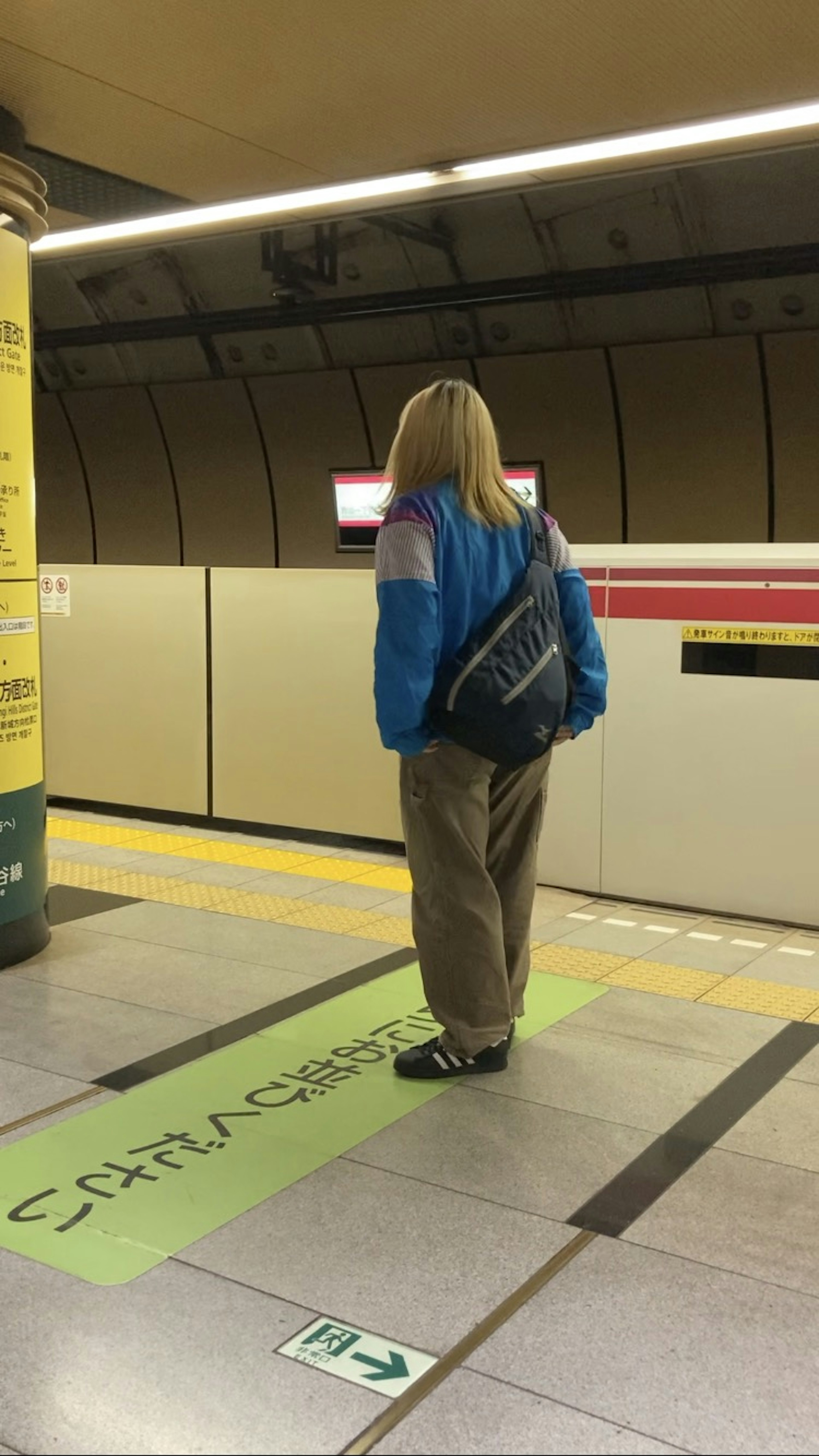 A woman standing on a subway platform wearing a blue jacket and beige pants near a green safety line