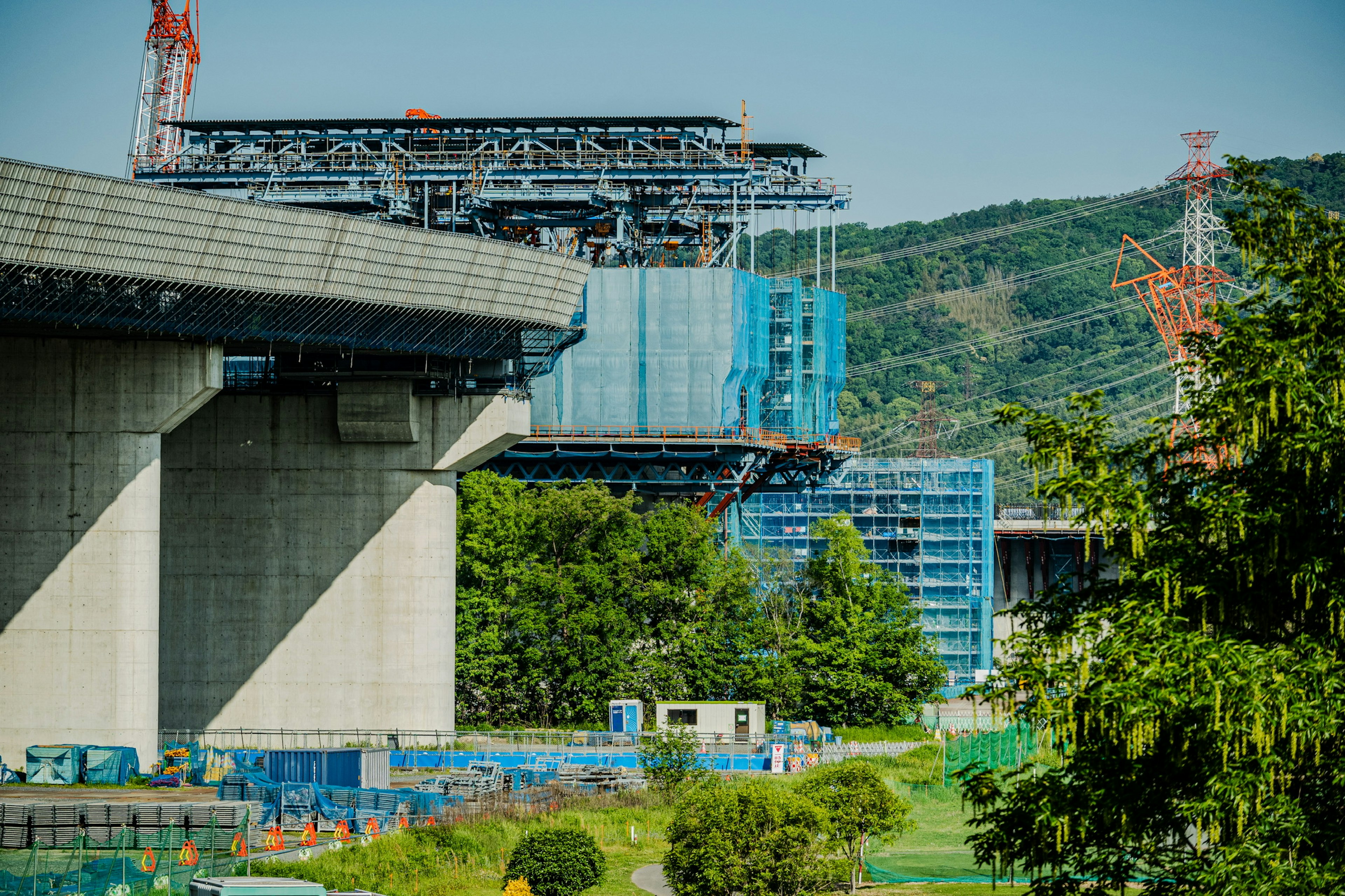 Under construction elevated bridge with surrounding construction site featuring lush greenery and cranes