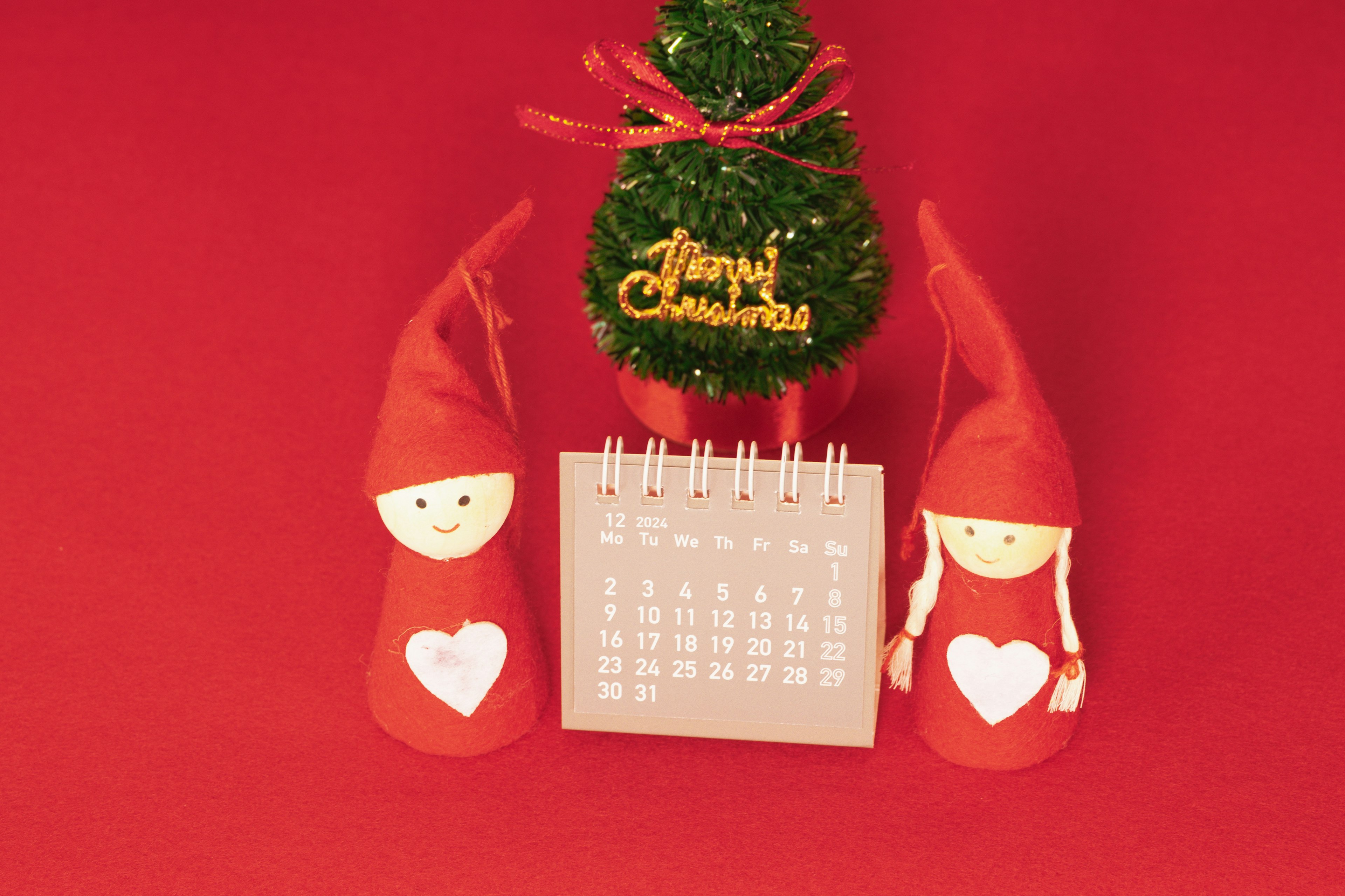 Two red-hatted dolls with hearts beside a calendar and a small Christmas tree on a red background