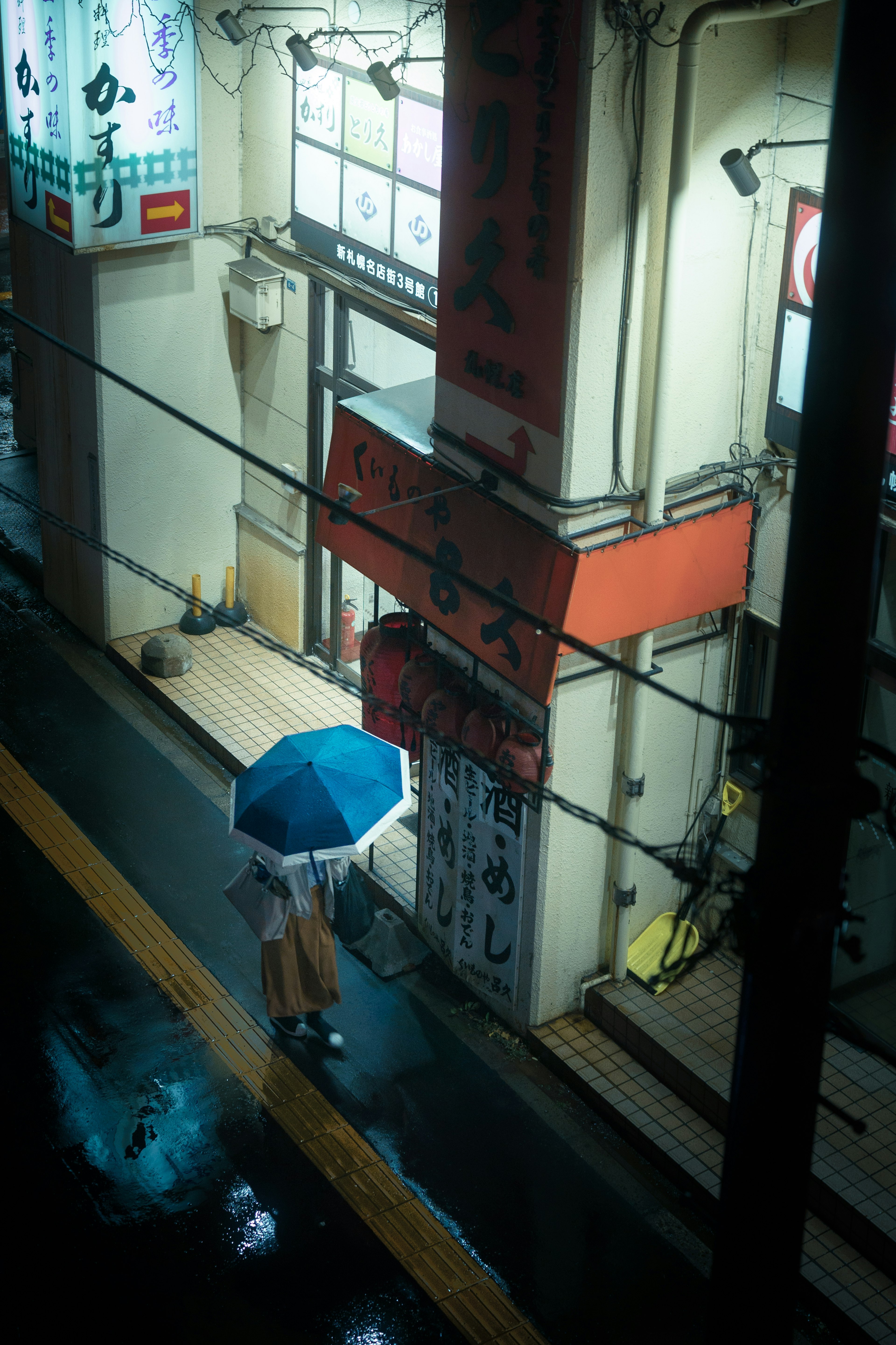 雨の中、青い傘を持った人物が街を歩いている夜のシーン