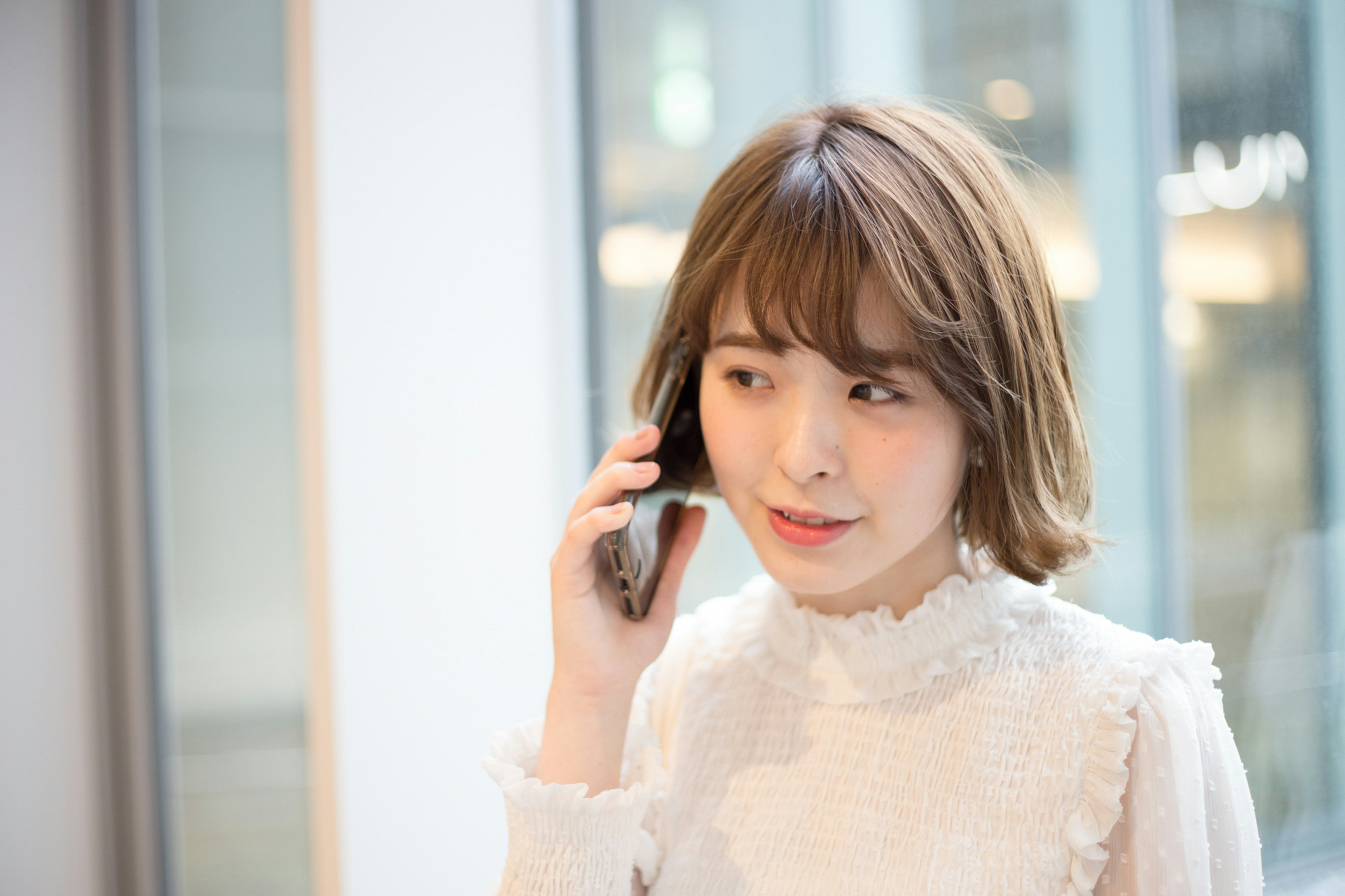 Portrait d'une femme au téléphone portant un chemisier blanc avec une coiffure douce et un arrière-plan de fenêtre en verre