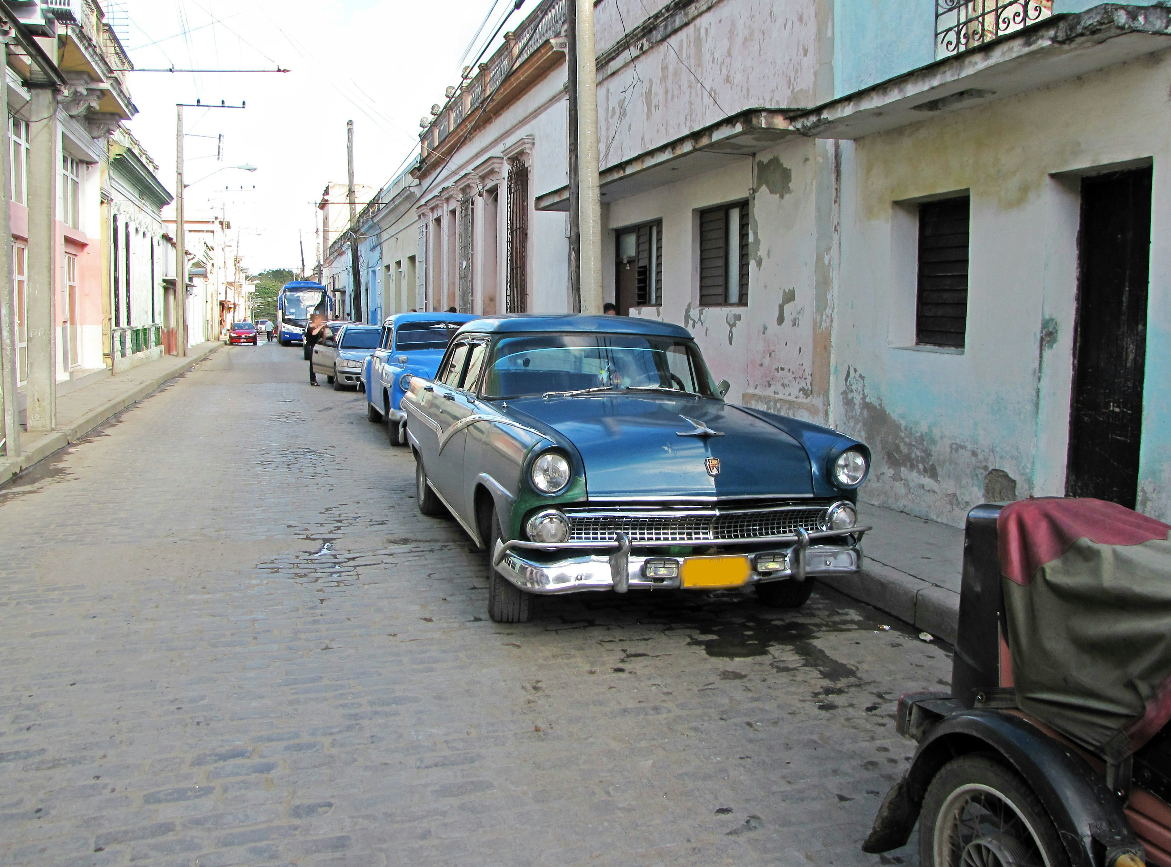 Mobil biru vintage diparkir di jalan berbatu dengan rumah-rumah berwarna