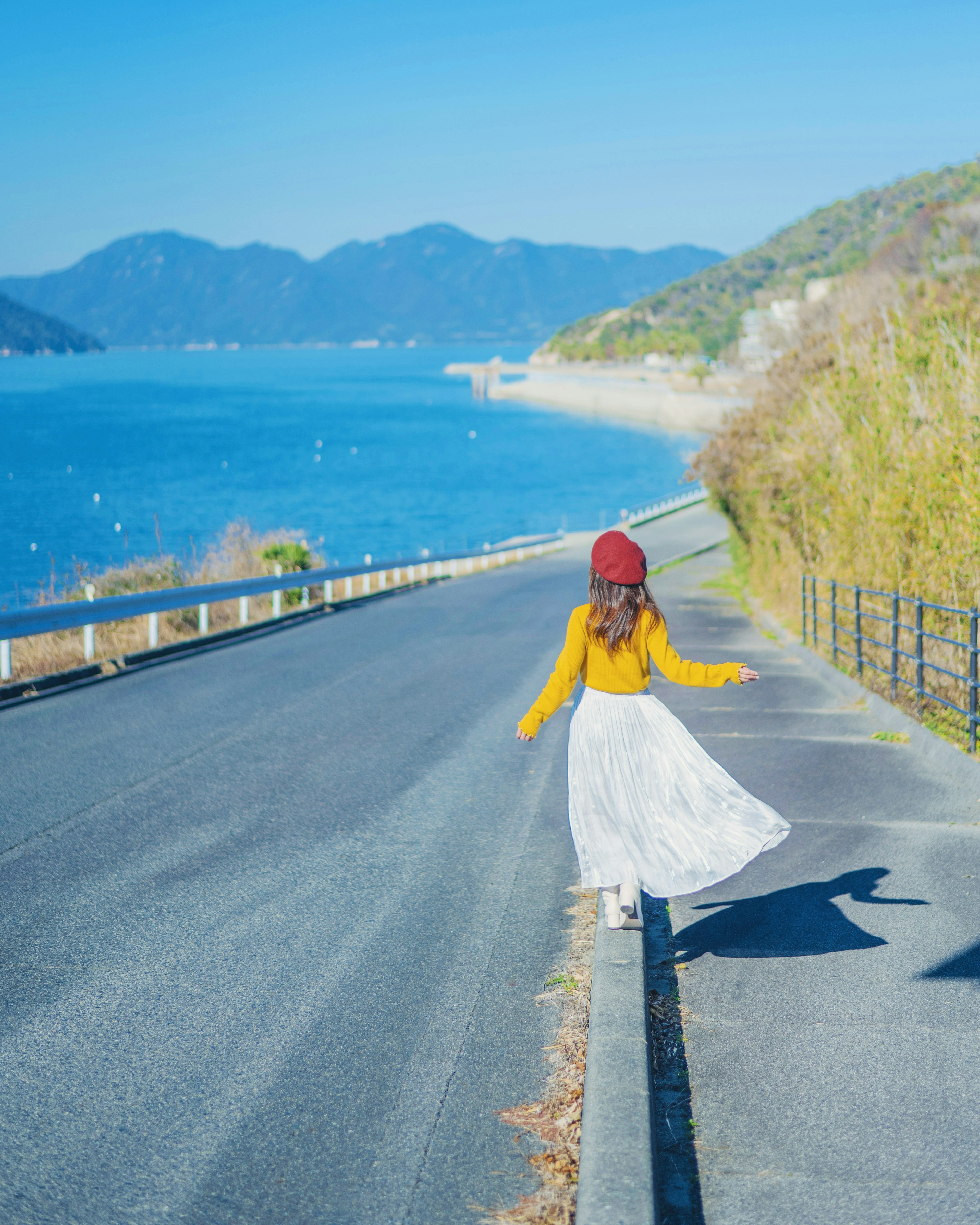 一位穿着黄色上衣和白色裙子的女性在蓝色海洋和山脉旁的道路上行走