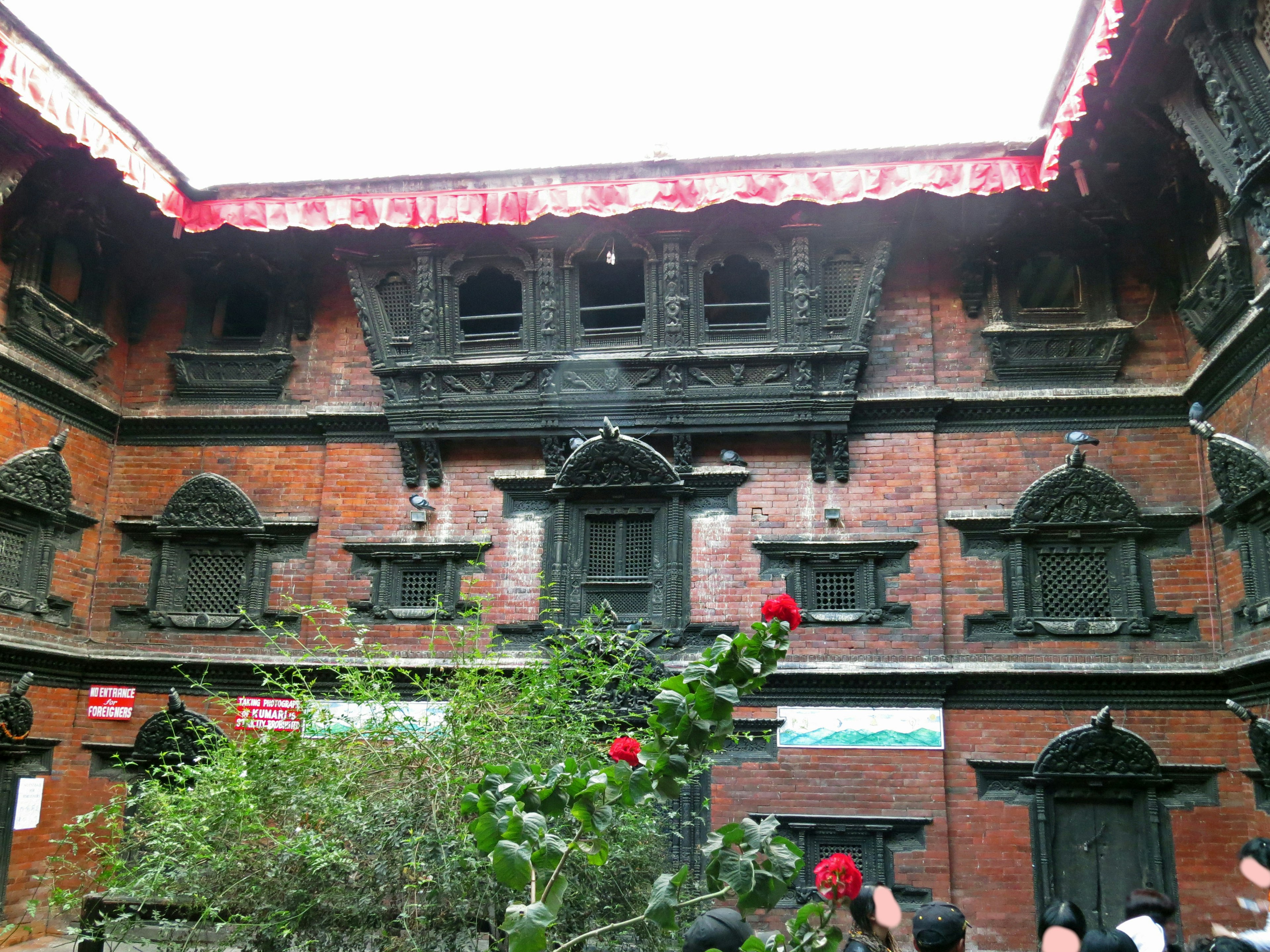 Arquitectura tradicional nepalí con ventanas decorativas en un patio con techos rojos y vegetación