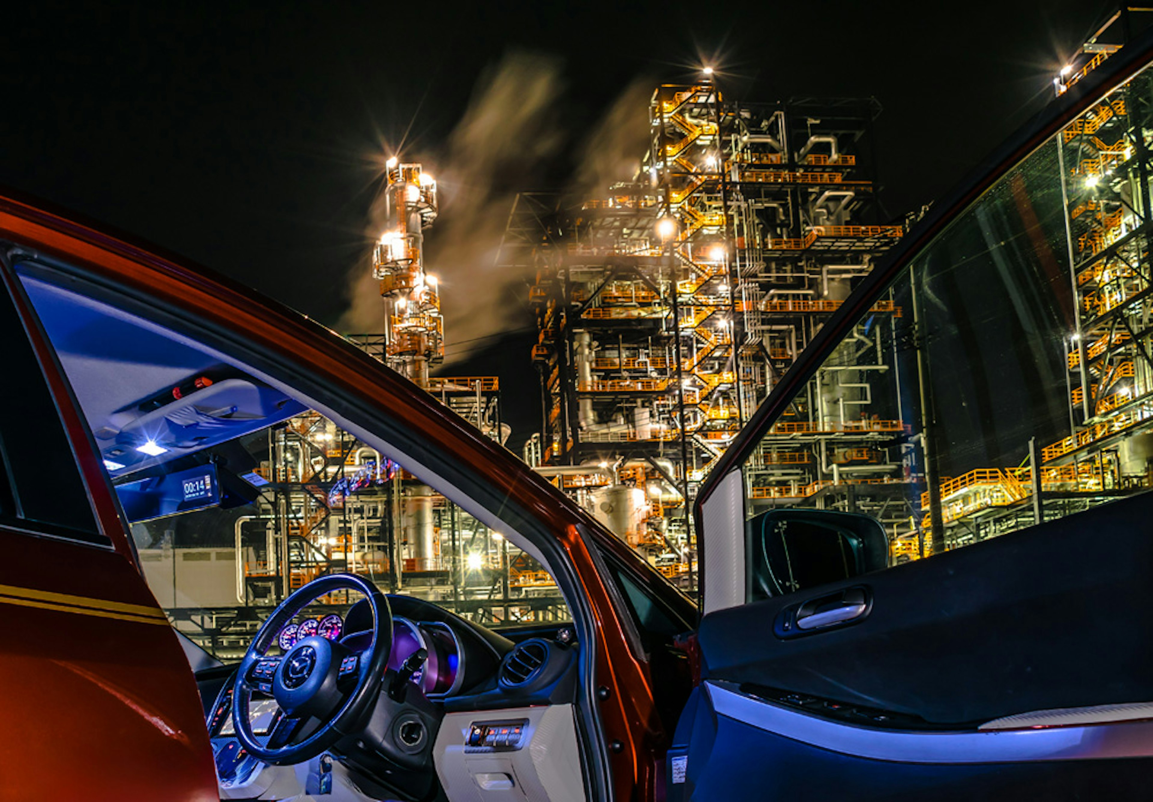 Interno di un'auto rossa con porta aperta che mostra una fabbrica industriale di notte