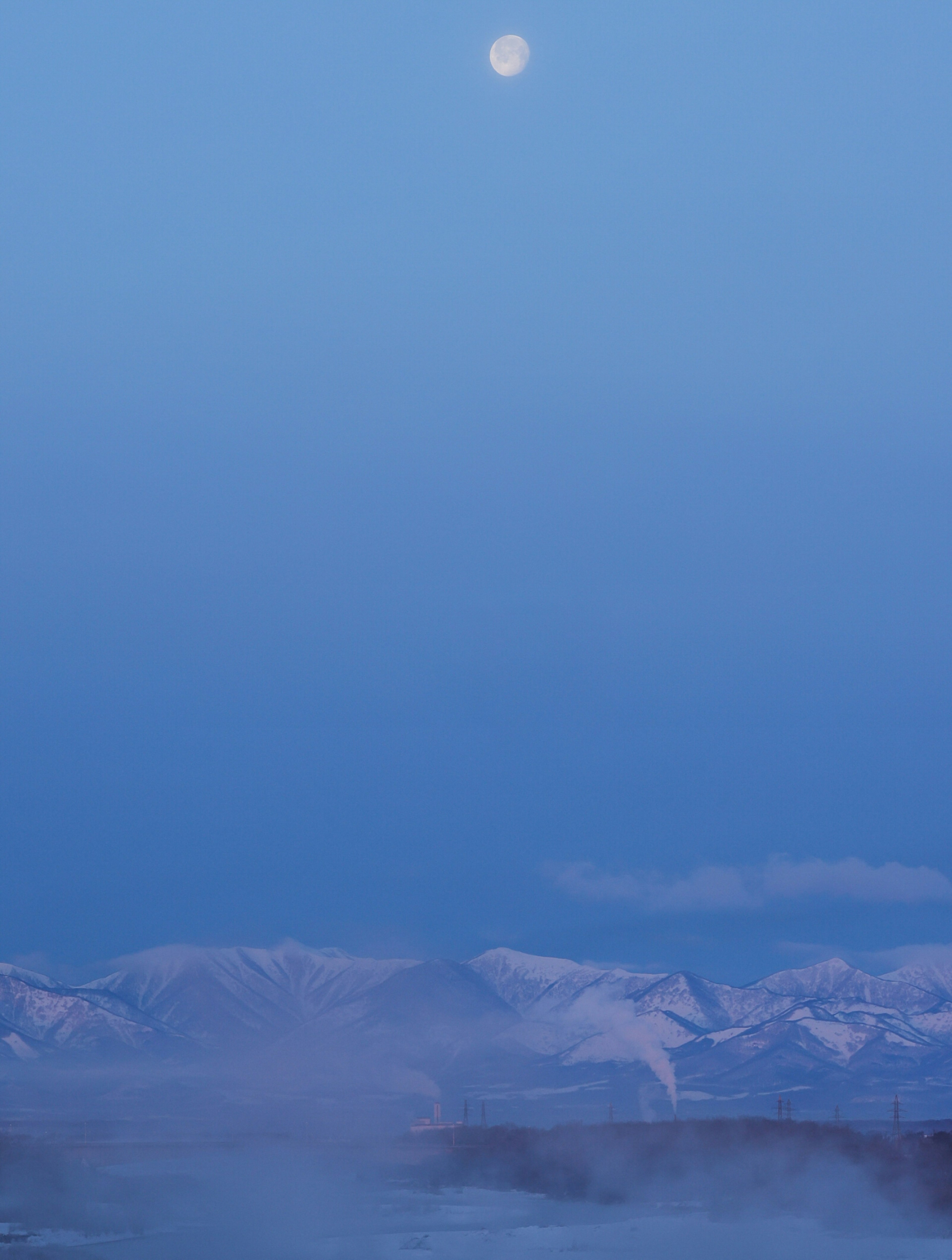 青い空の下に浮かぶ月と雪をかぶった山々