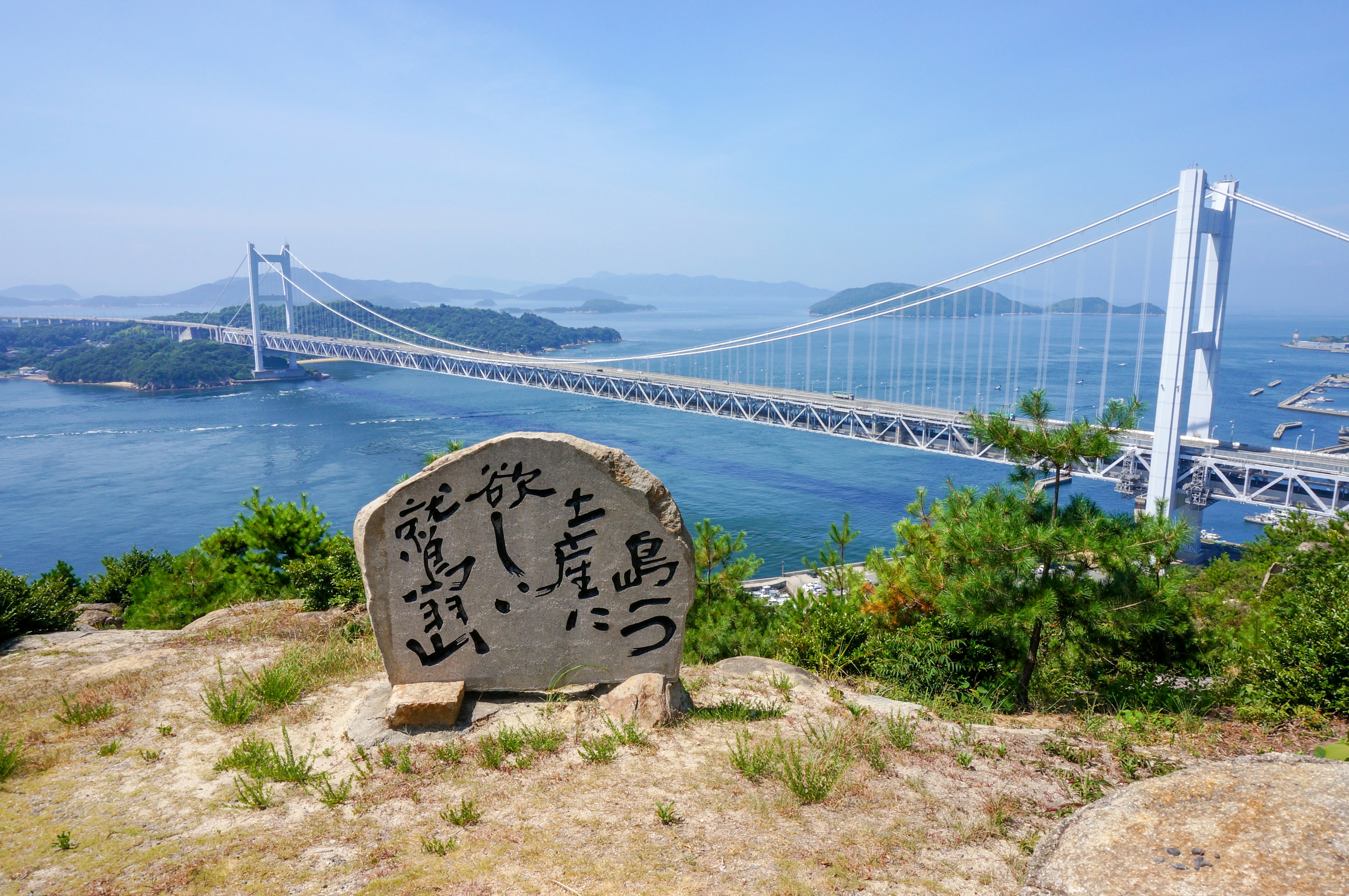 Steinmonument mit japanischer Inschrift mit Blick auf ein blaues Meer und eine Brücke