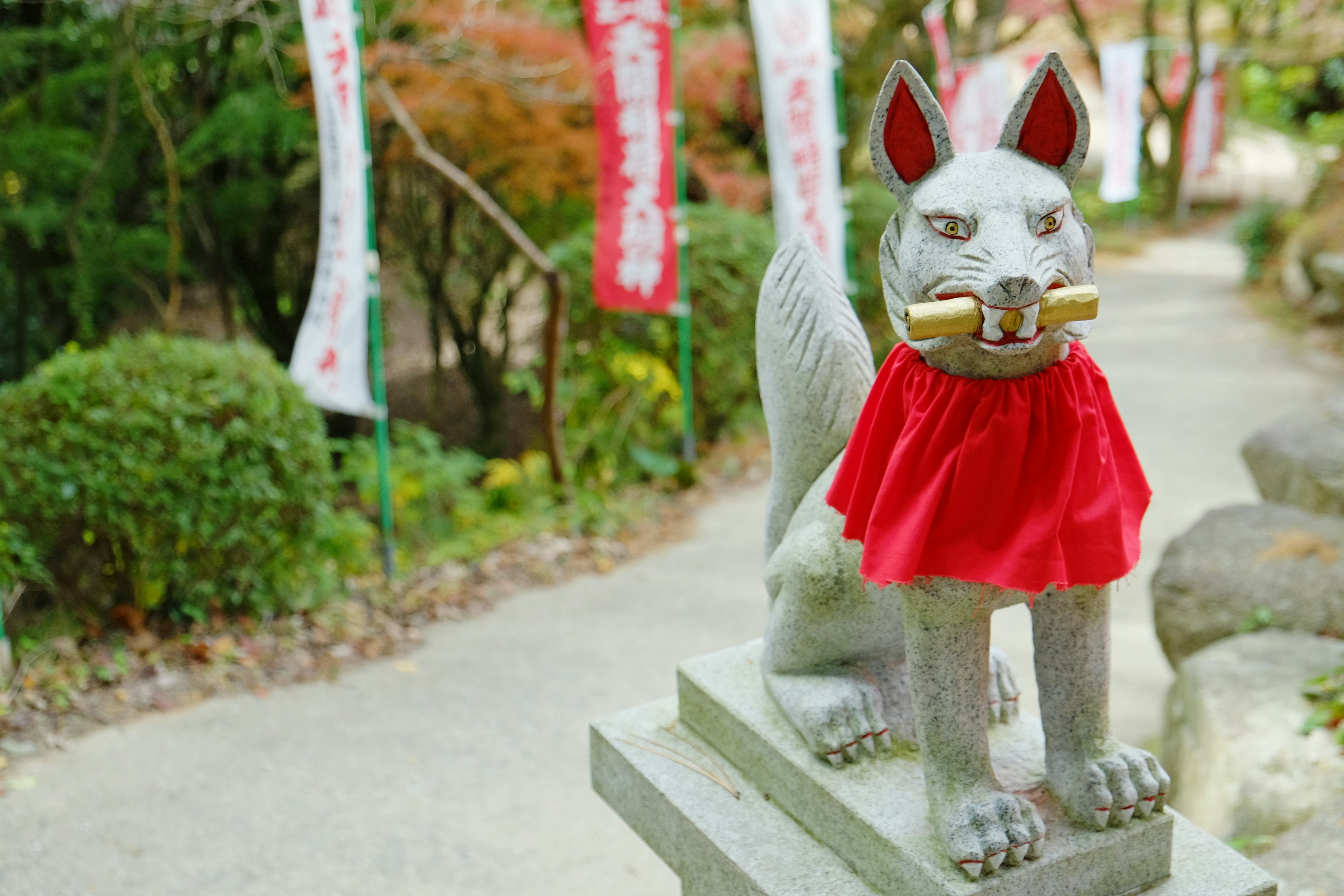 Eine Fuchsstatue mit rotem Umhang steht neben einem Weg mit Bäumen und Bannern im Hintergrund