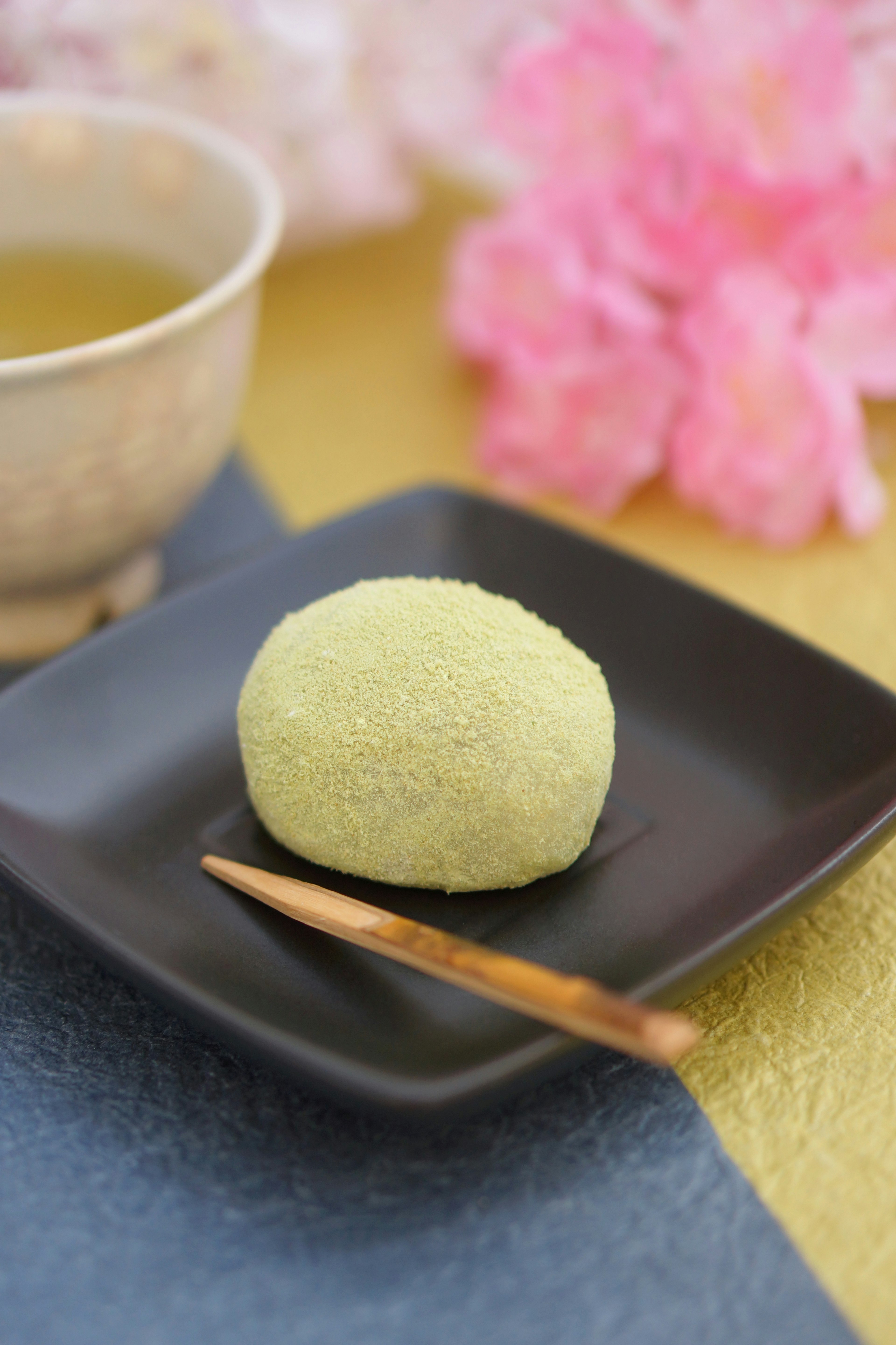 Matcha mochi on a black plate with a wooden stick beside a cup of green tea and cherry blossoms in the background