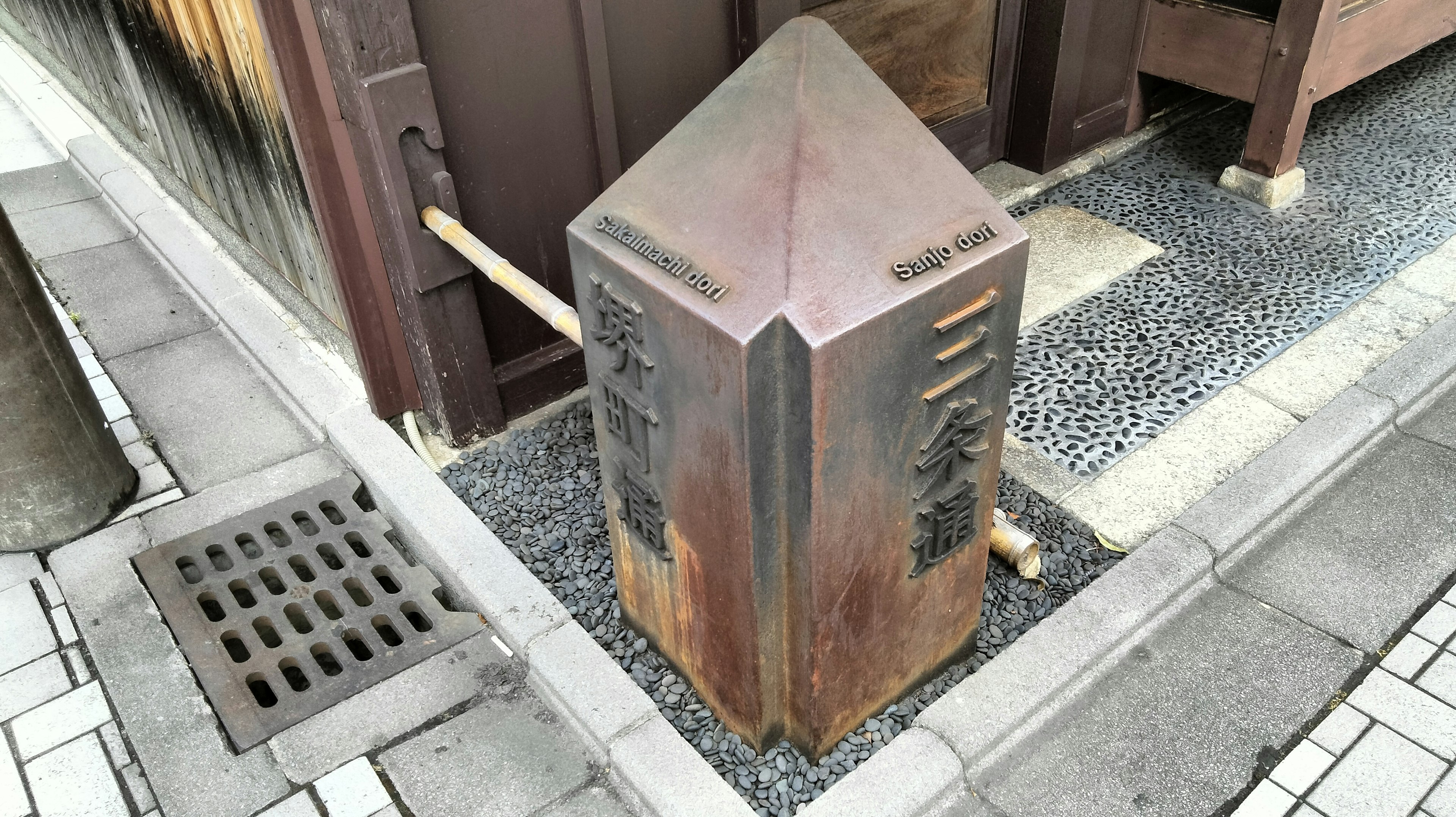 A vintage metal corner post stands at a street corner surrounded by stone pavement and wooden structures