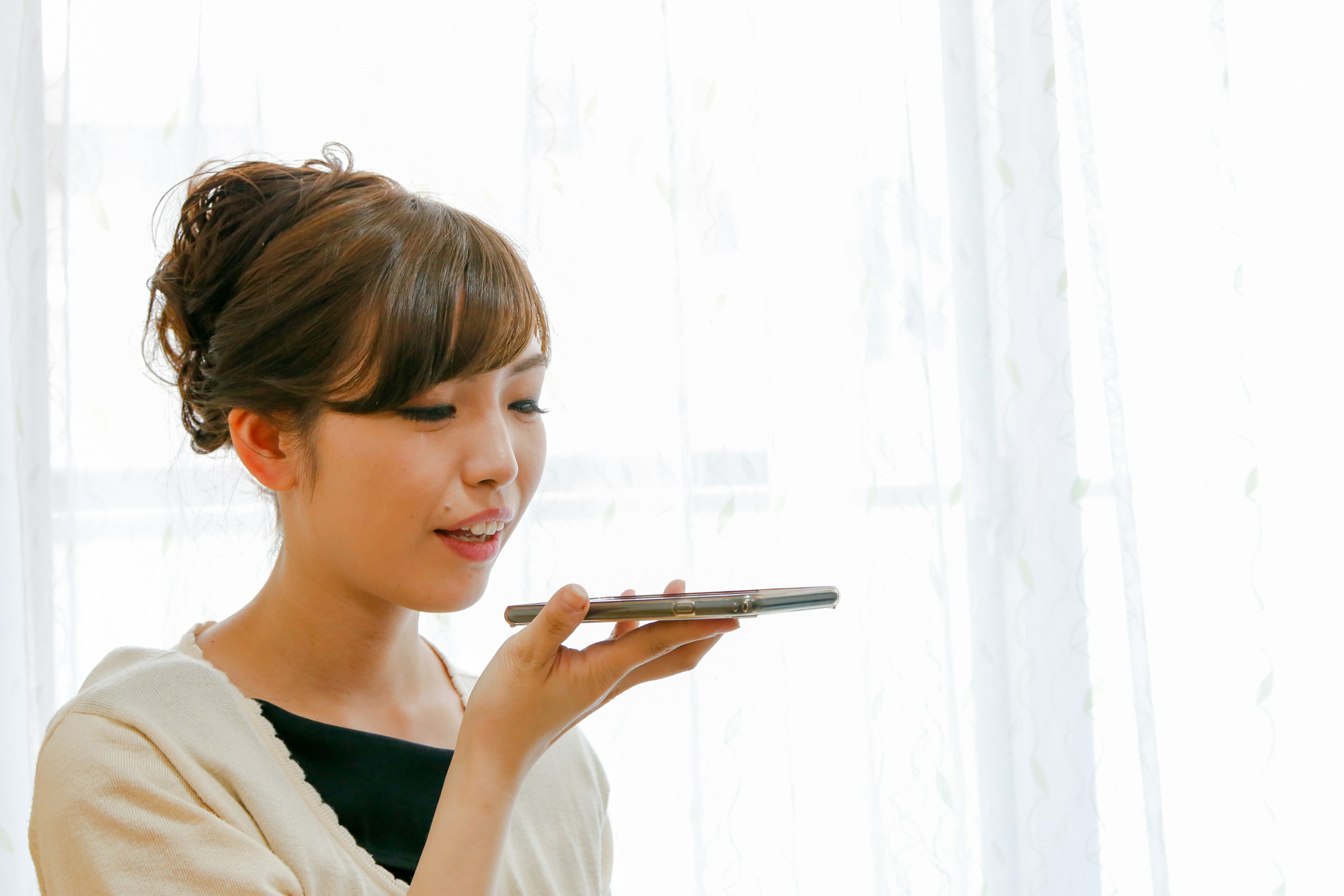 Femme envoyant un message vocal sur un smartphone souriant devant une fenêtre lumineuse