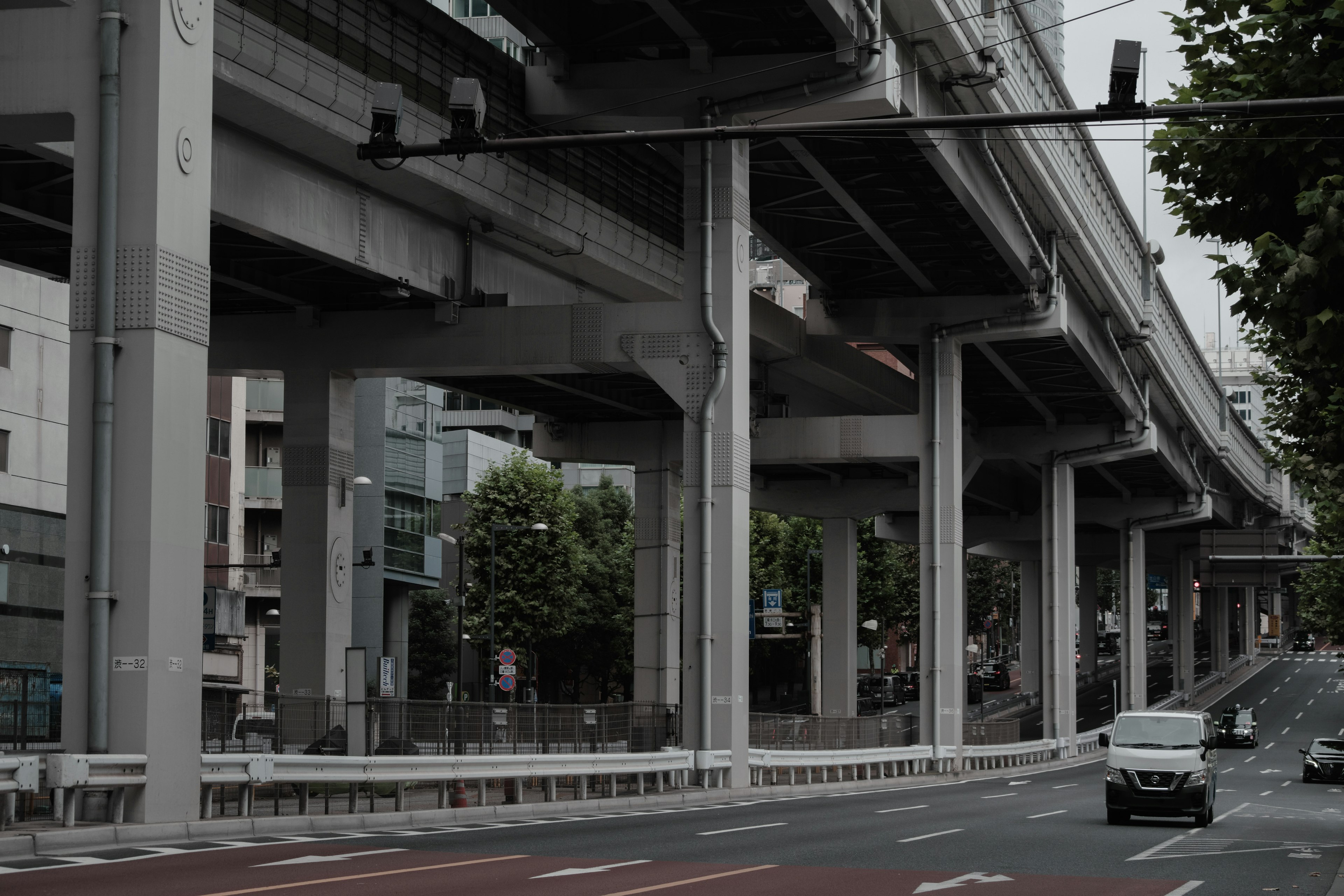 Urban scene under an elevated highway featuring concrete pillars and green trees