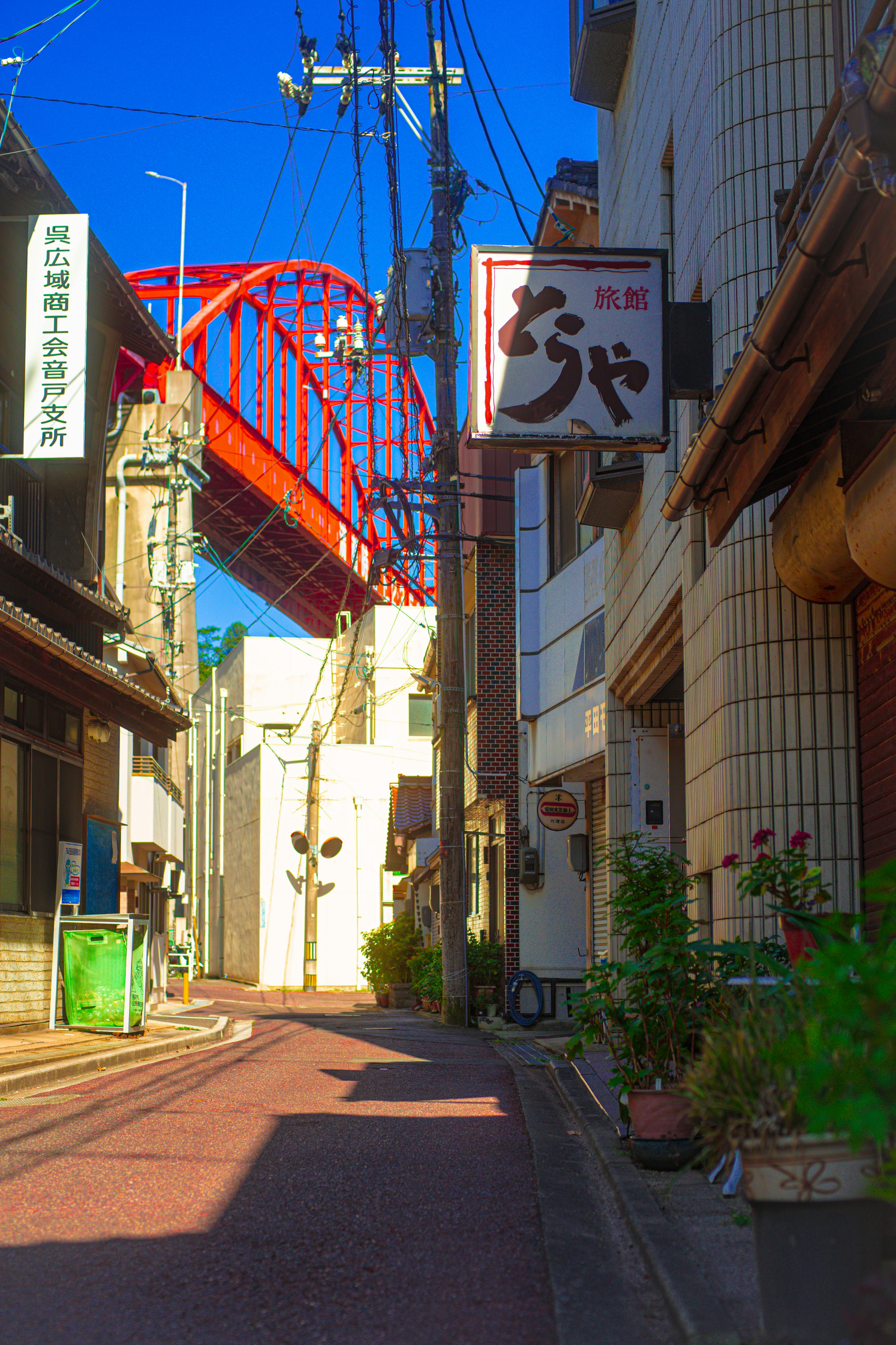 青空の下の狭い路地にある飲食店の看板と赤い橋が見える