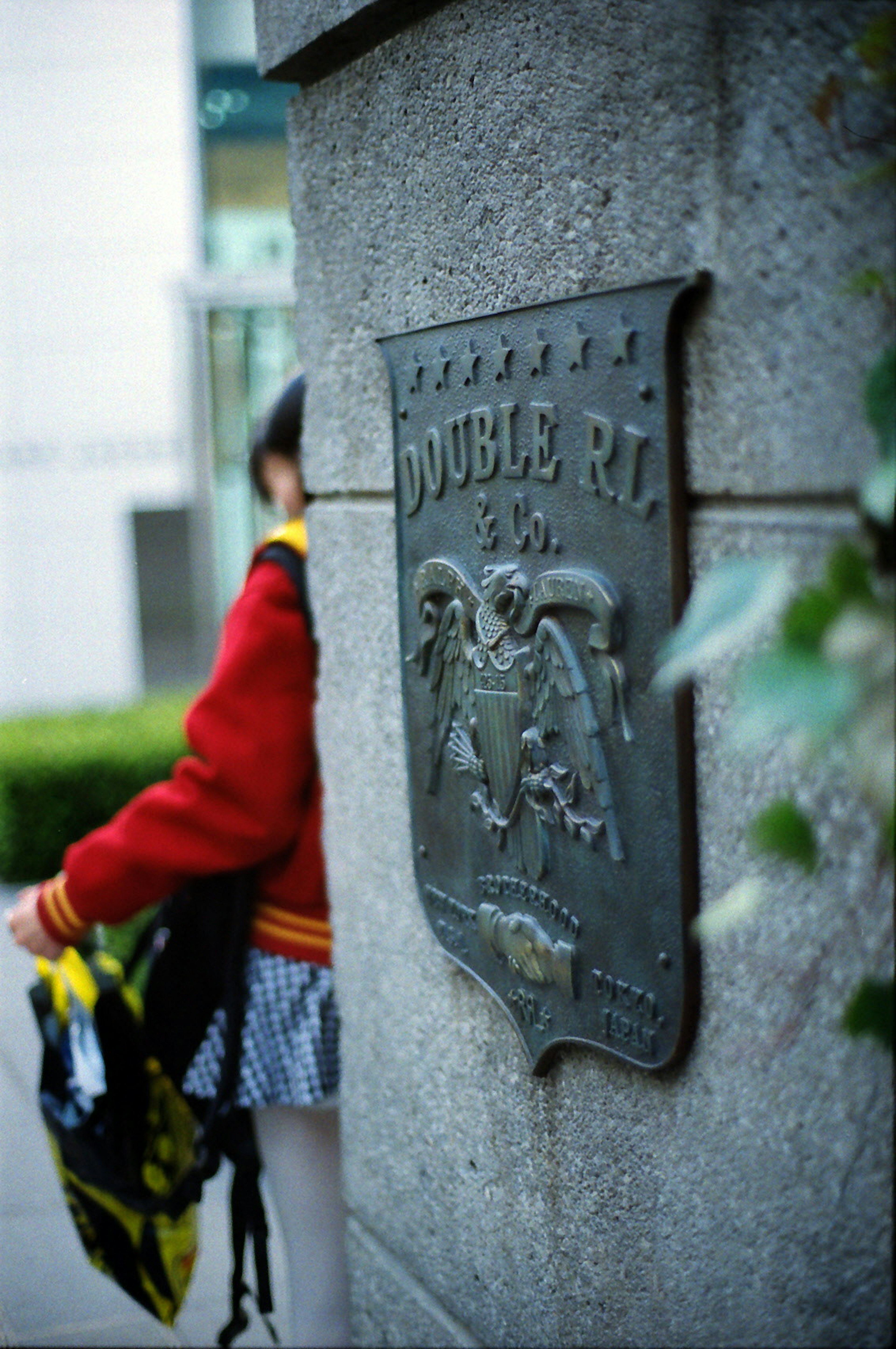 A person in a red jacket stands near a stone pillar featuring a metal plaque with an embossed emblem