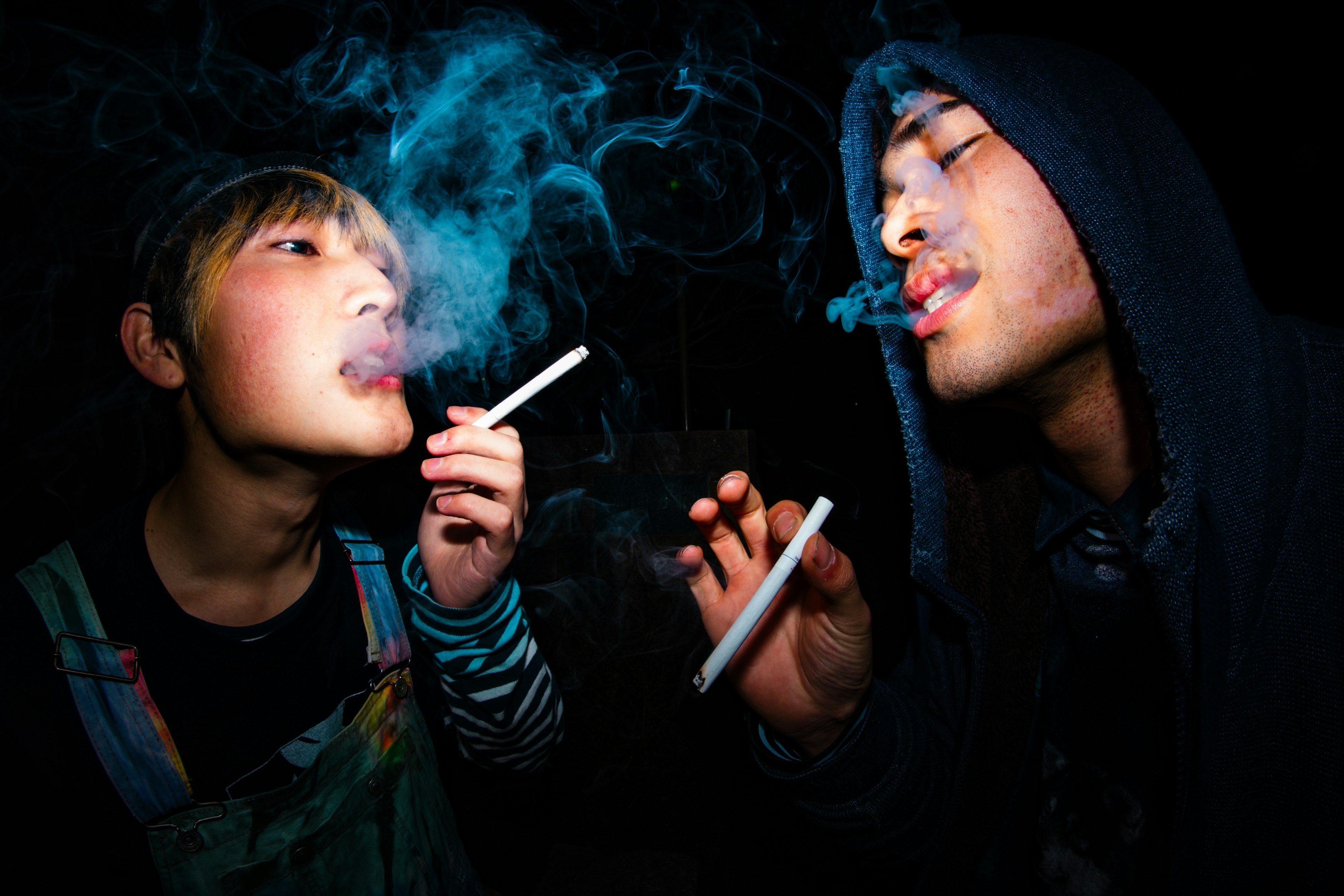 Photo of two young individuals smoking with a dark background and blue smoke