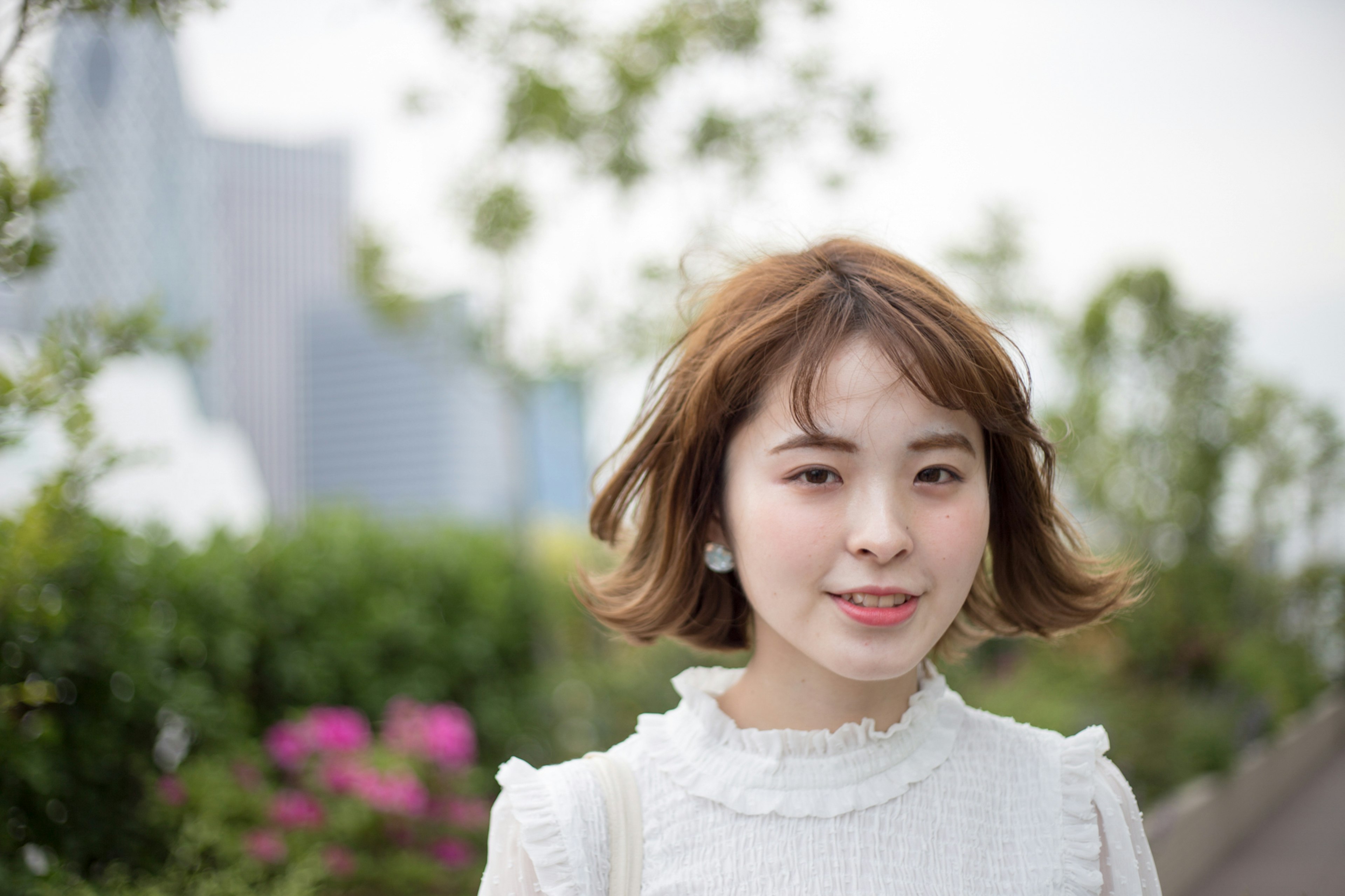 Femme souriante dans un parc portant un chemisier blanc cheveux courts plantes vertes et fleurs en arrière-plan paysage urbain flou