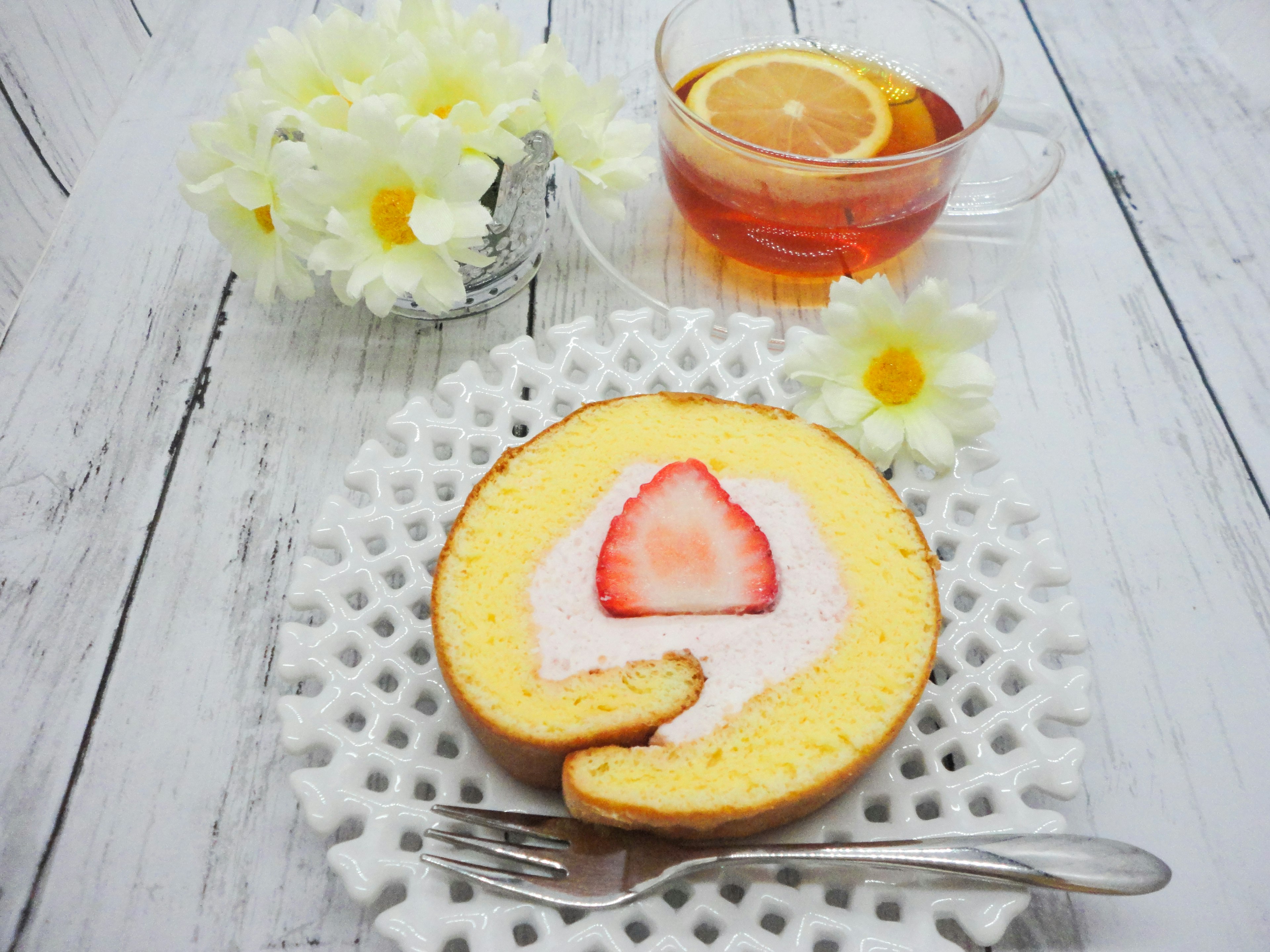 Pastel enrollado amarillo con fresa encima dispuesto junto a flores blancas y té