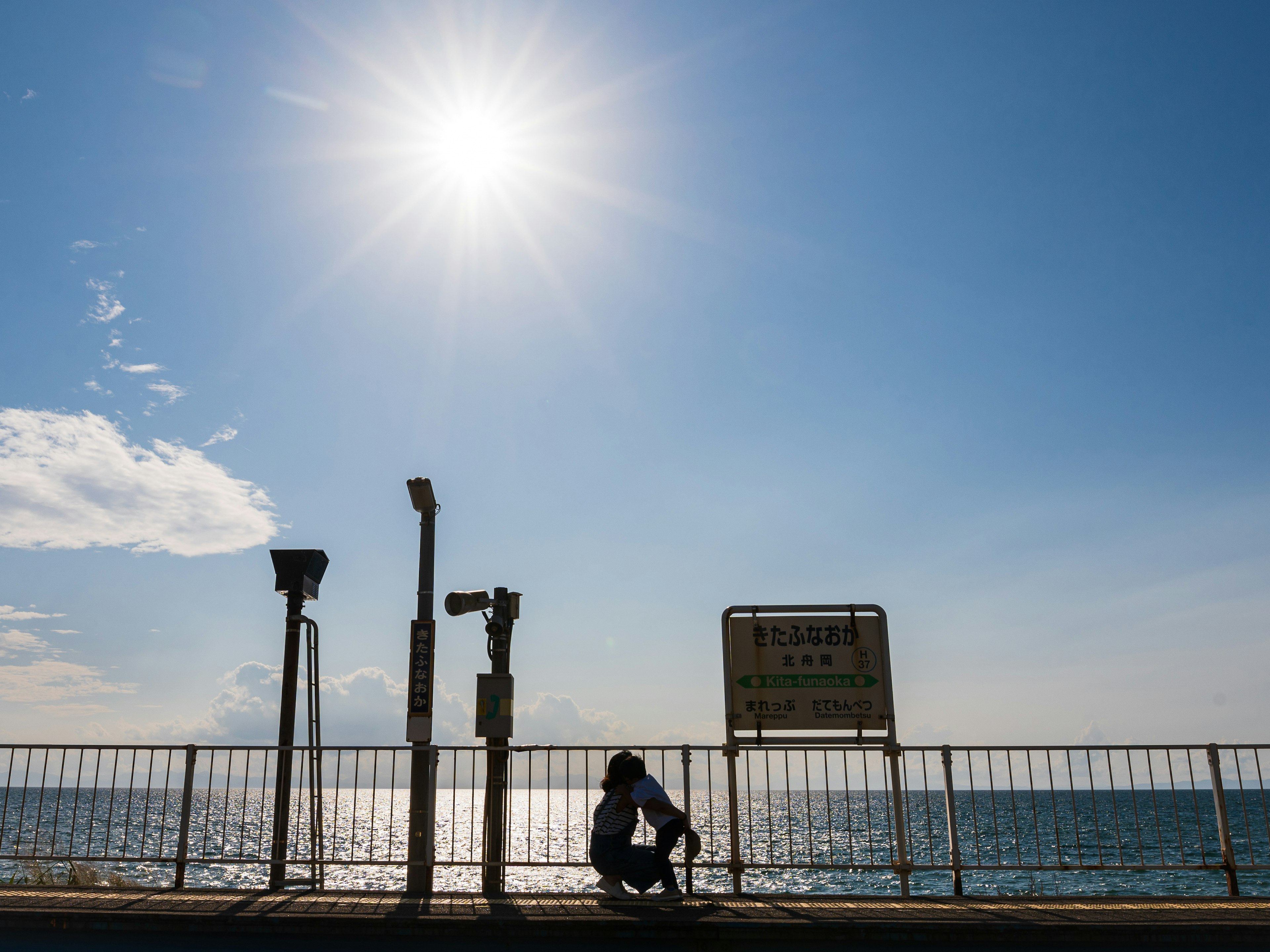 A silhouette of a person leaning against a fence under a bright sun and blue sea