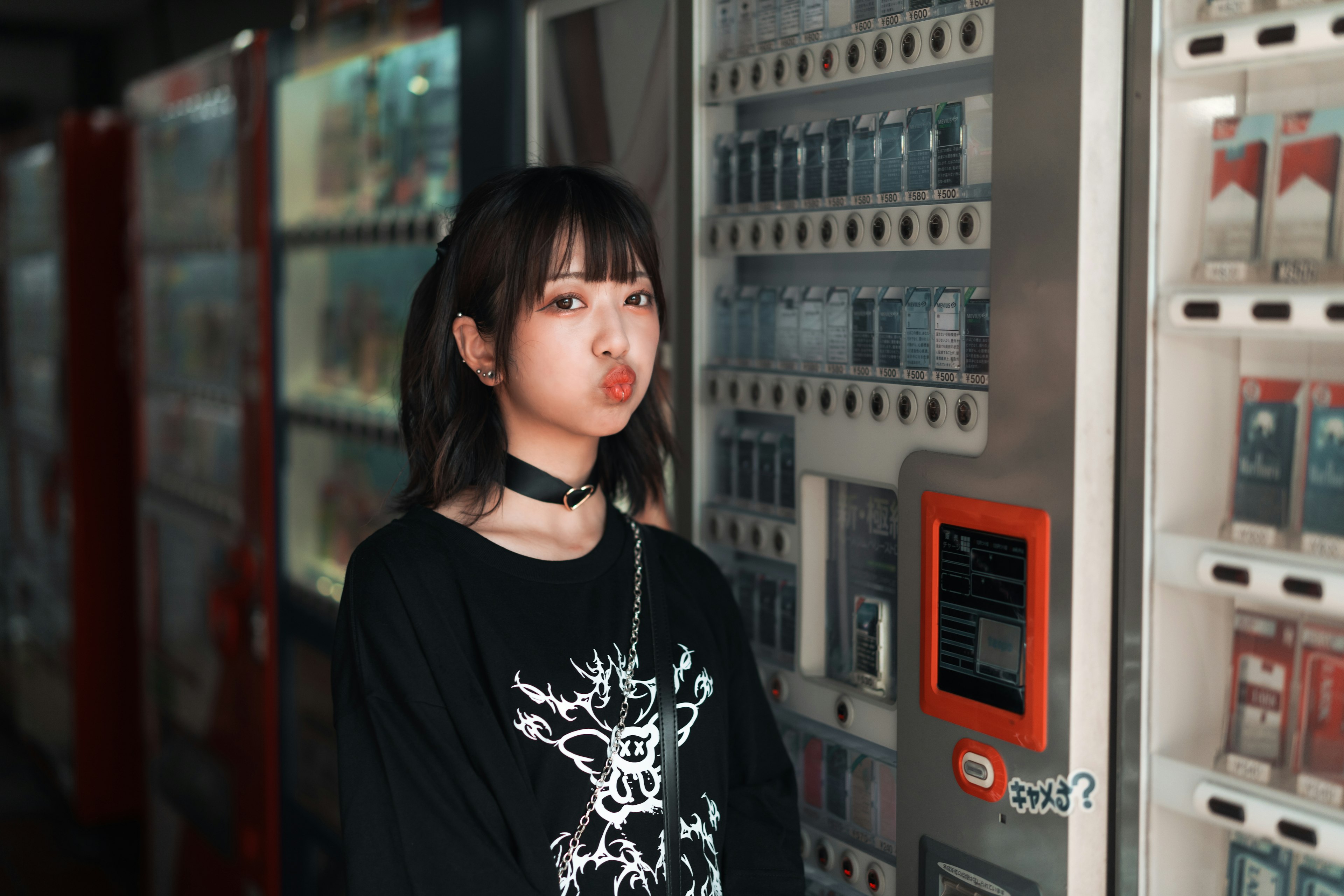 自動販売機の前に立つ女性のポートレート 黒いTシャツとチョーカーを着用 髪は肩までの長さ