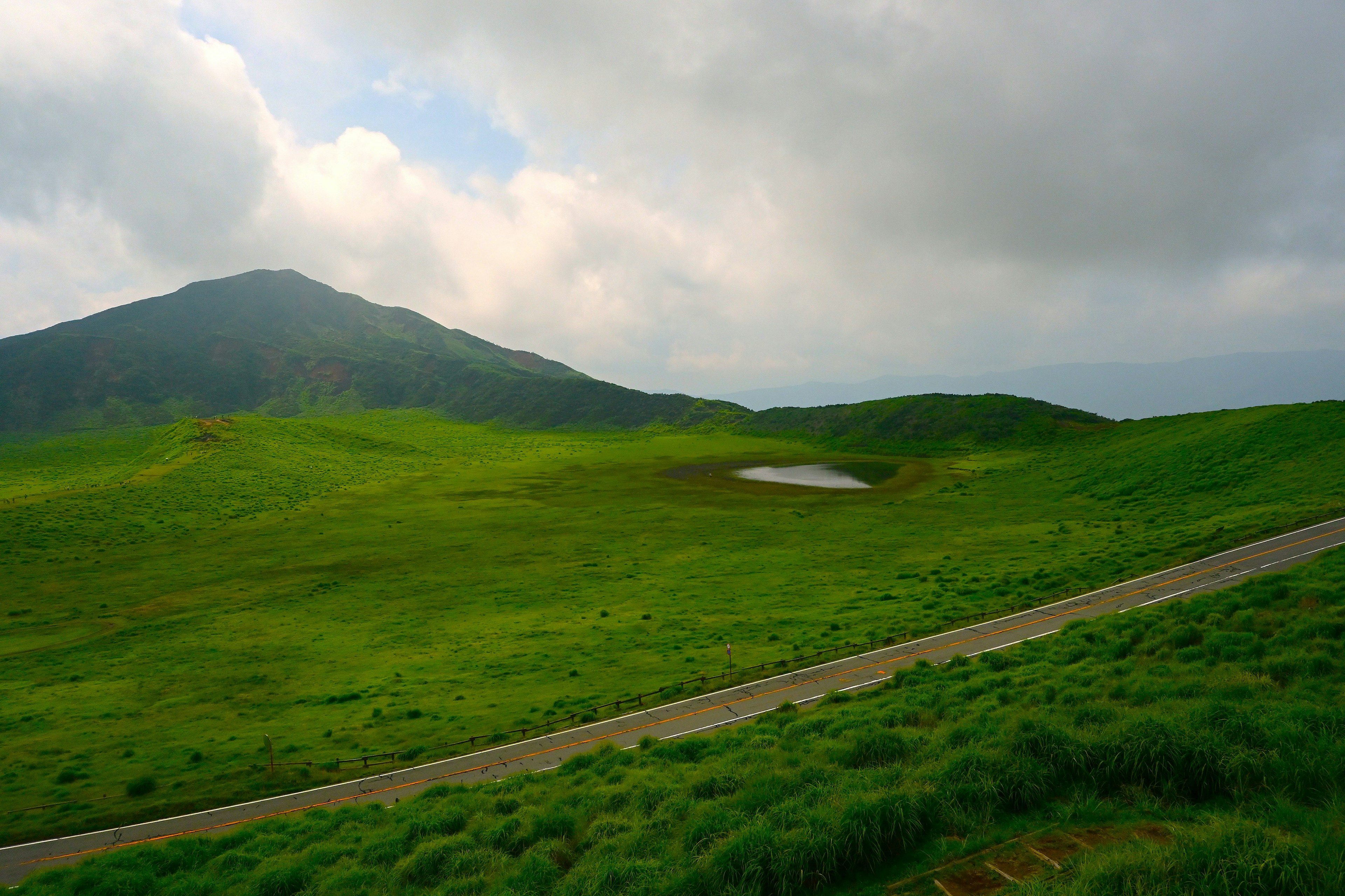 緑の丘と山が広がる風景に小さな池がある