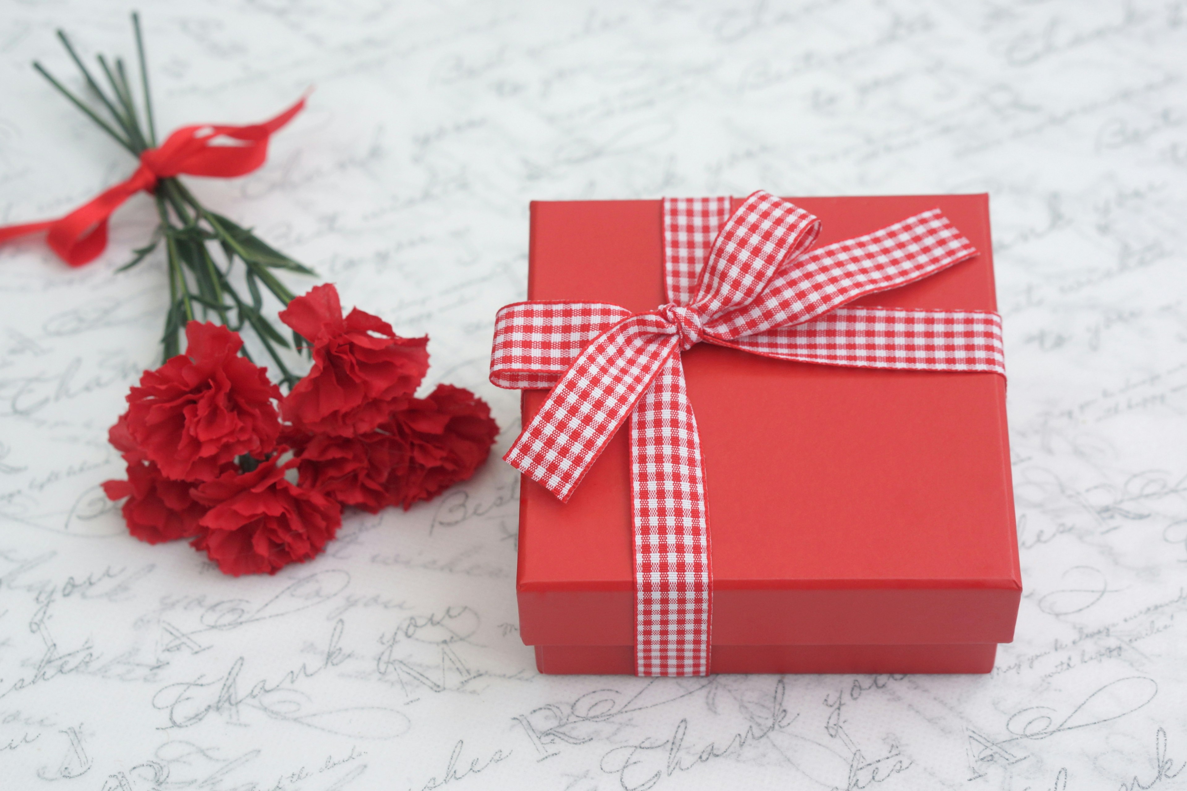 Red gift box with a checkered ribbon next to a bouquet of red carnations