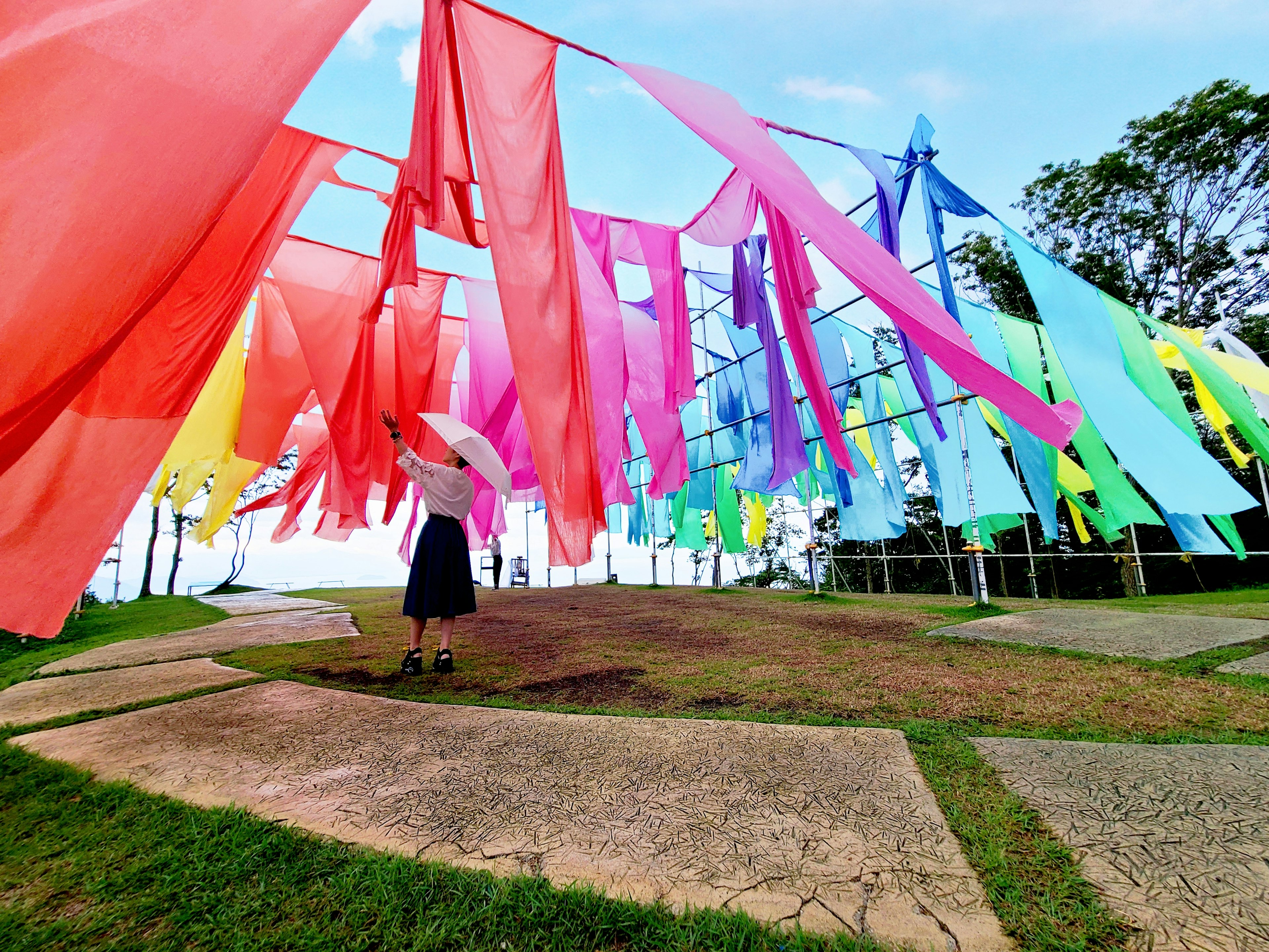 A woman holding an umbrella stands among colorful flowing fabric in an outdoor art installation