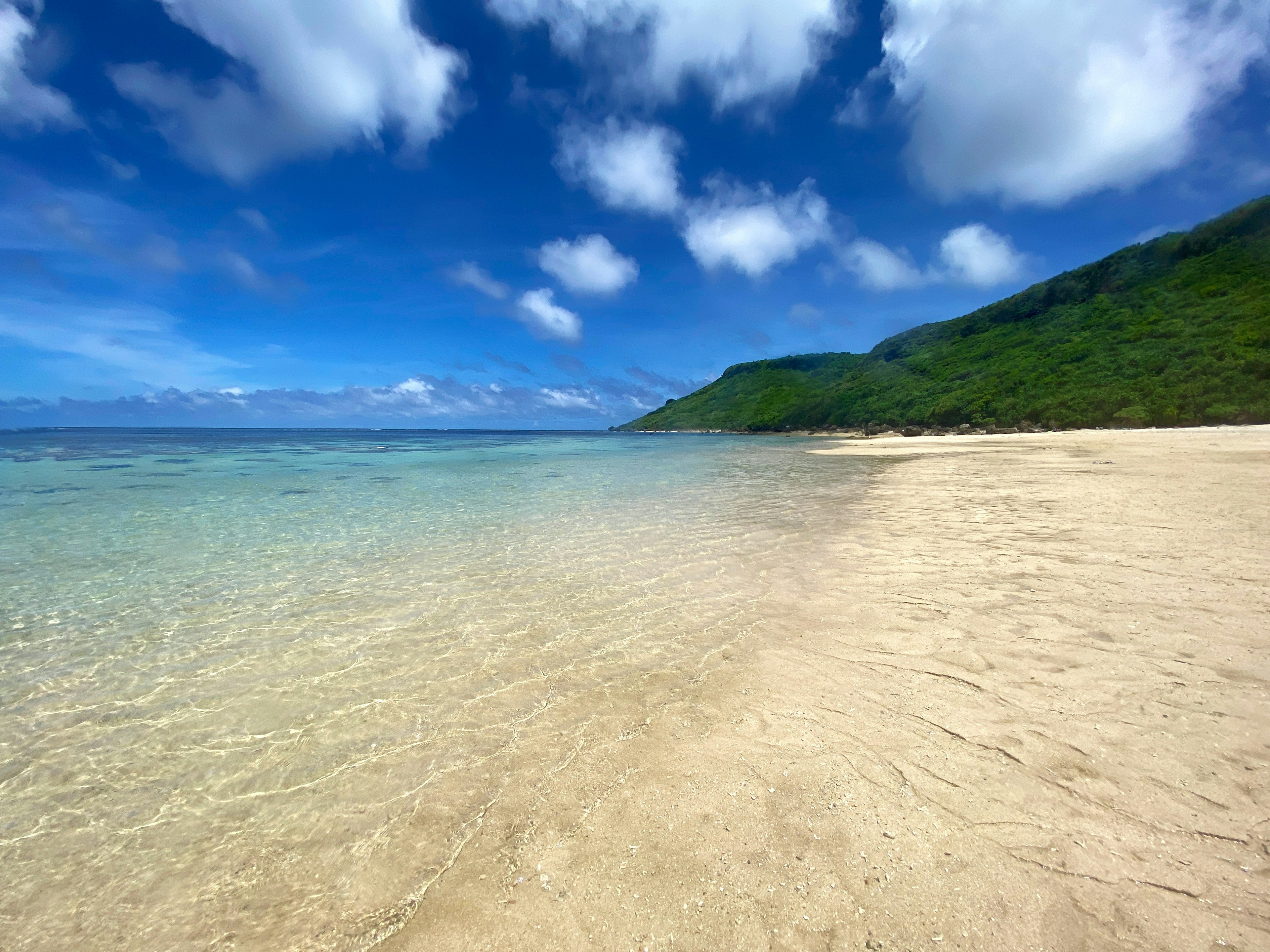 Strandszene mit blauem Himmel und weißen Wolken klares Wasser und sandiger Ufer grüne Hügel im Hintergrund