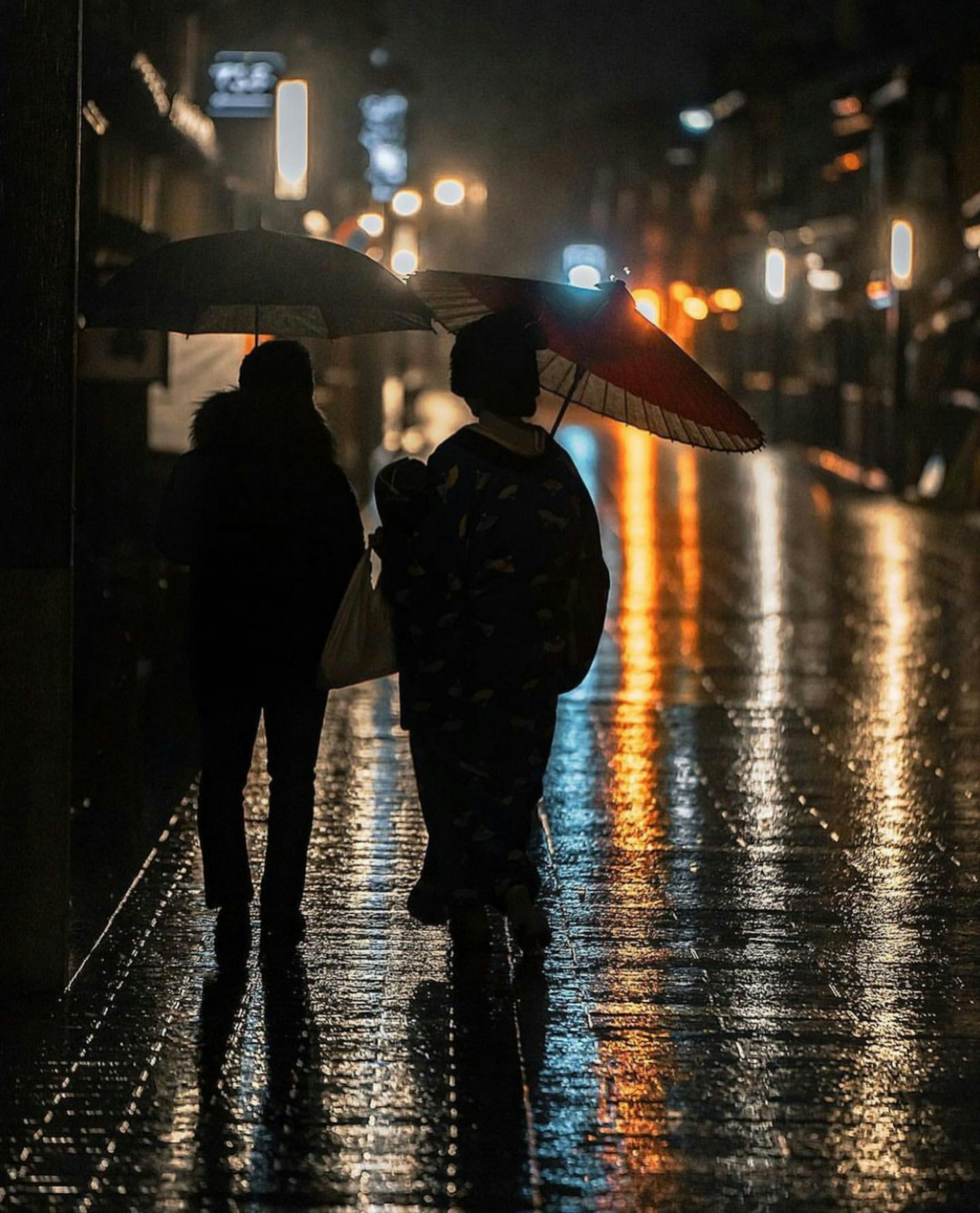 Silhouetten von zwei Personen, die bei Nacht unter Regenschirmen auf einer nassen Straße gehen