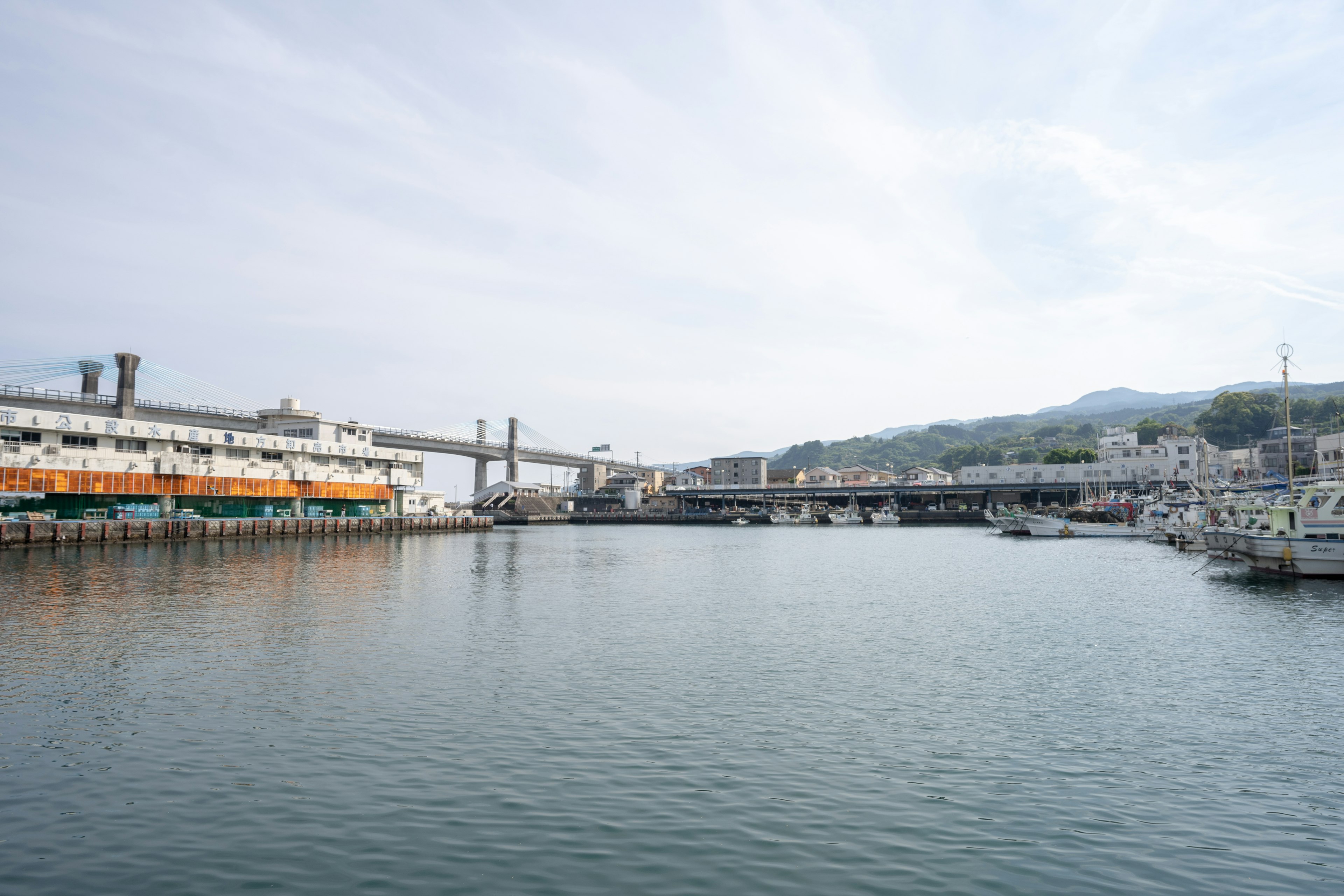 Escena de puerto con barcos y agua tranquila con un puente y montañas al fondo