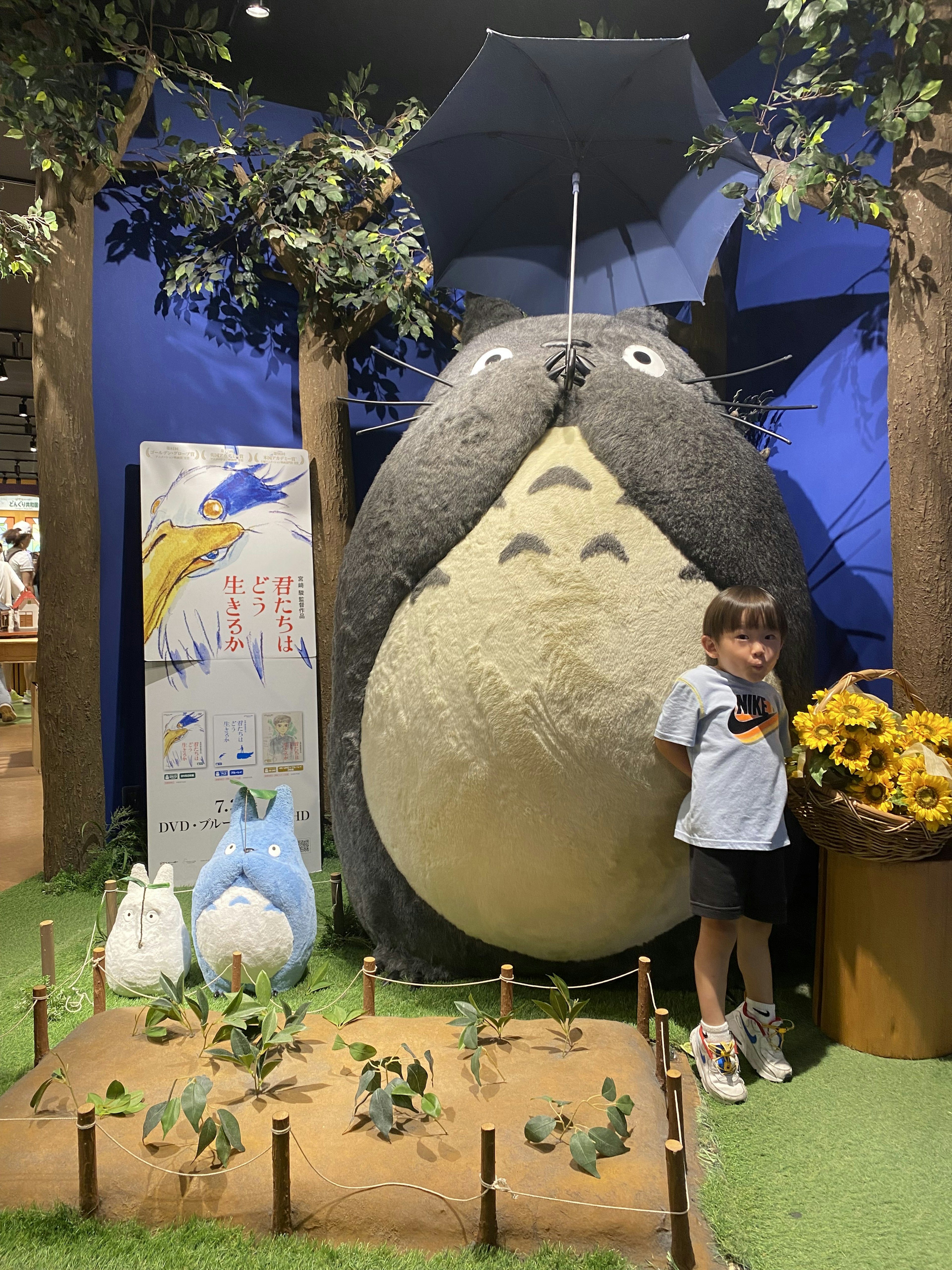 Một đứa trẻ đứng bên cạnh một con thú nhồi bông Totoro lớn, xung quanh là cây cối và cỏ xanh