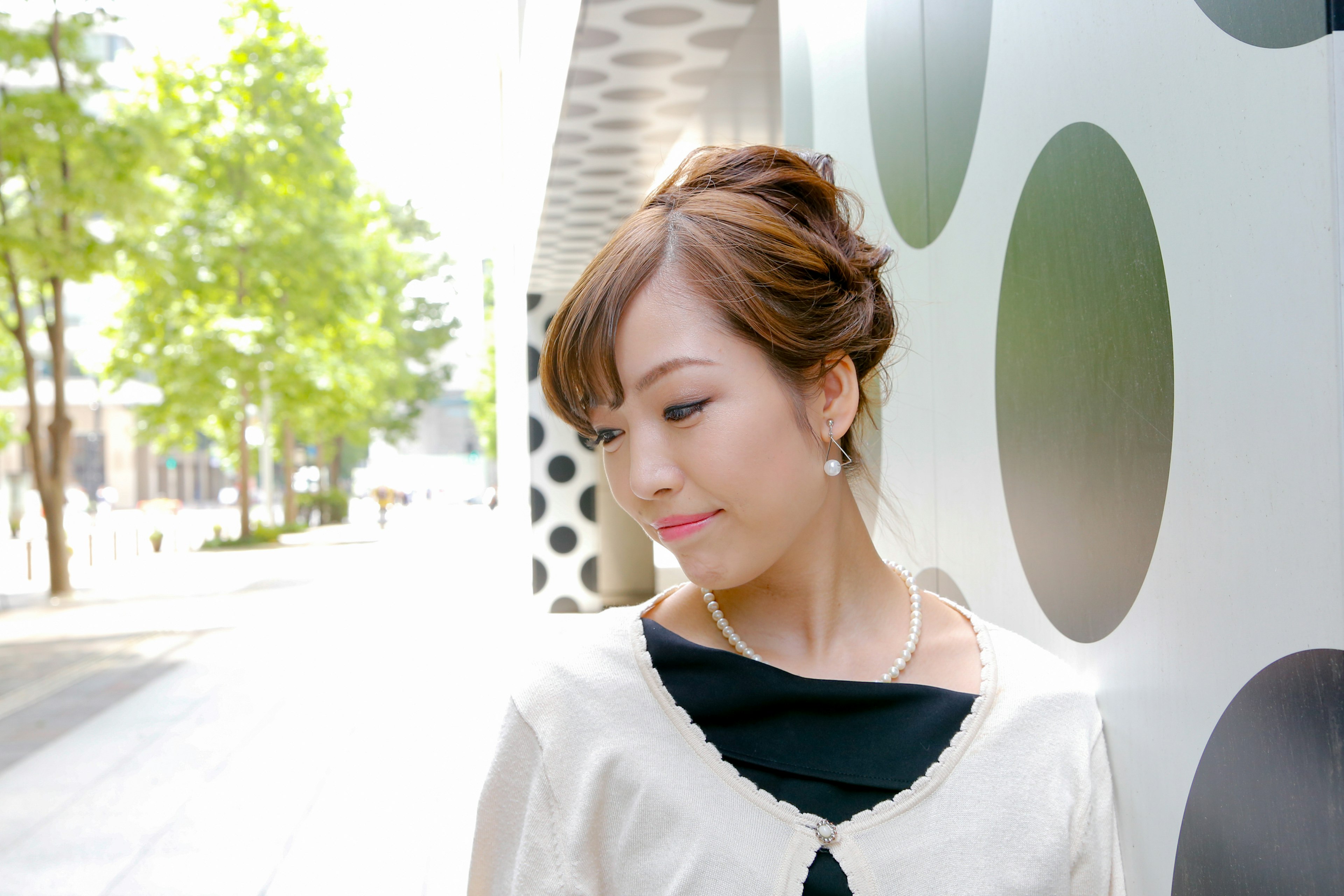 A woman wearing a black top and a white cardigan with a dotted wall and green trees in the background