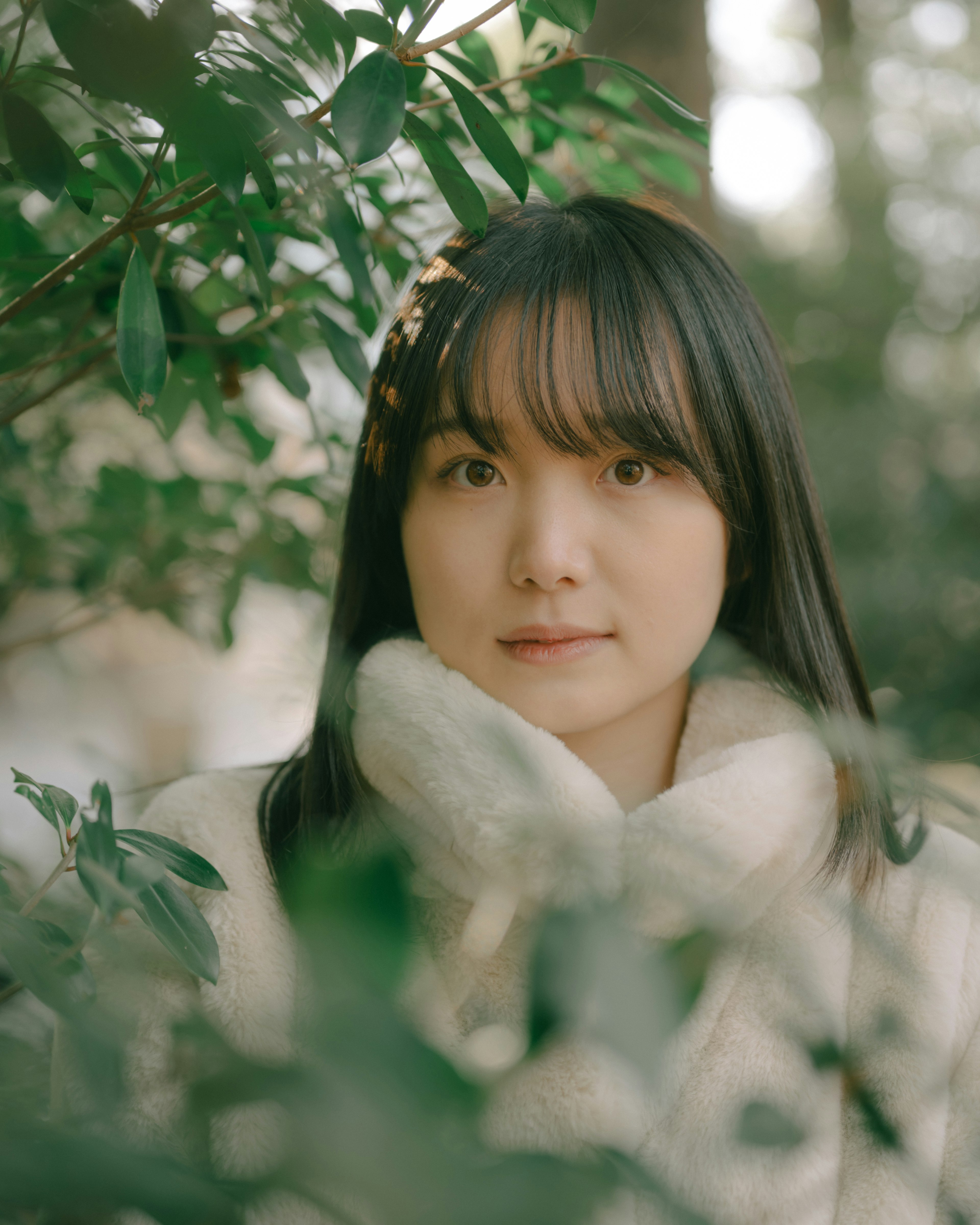 Portrait of a young woman surrounded by green leaves wearing a warm coat with a serene expression