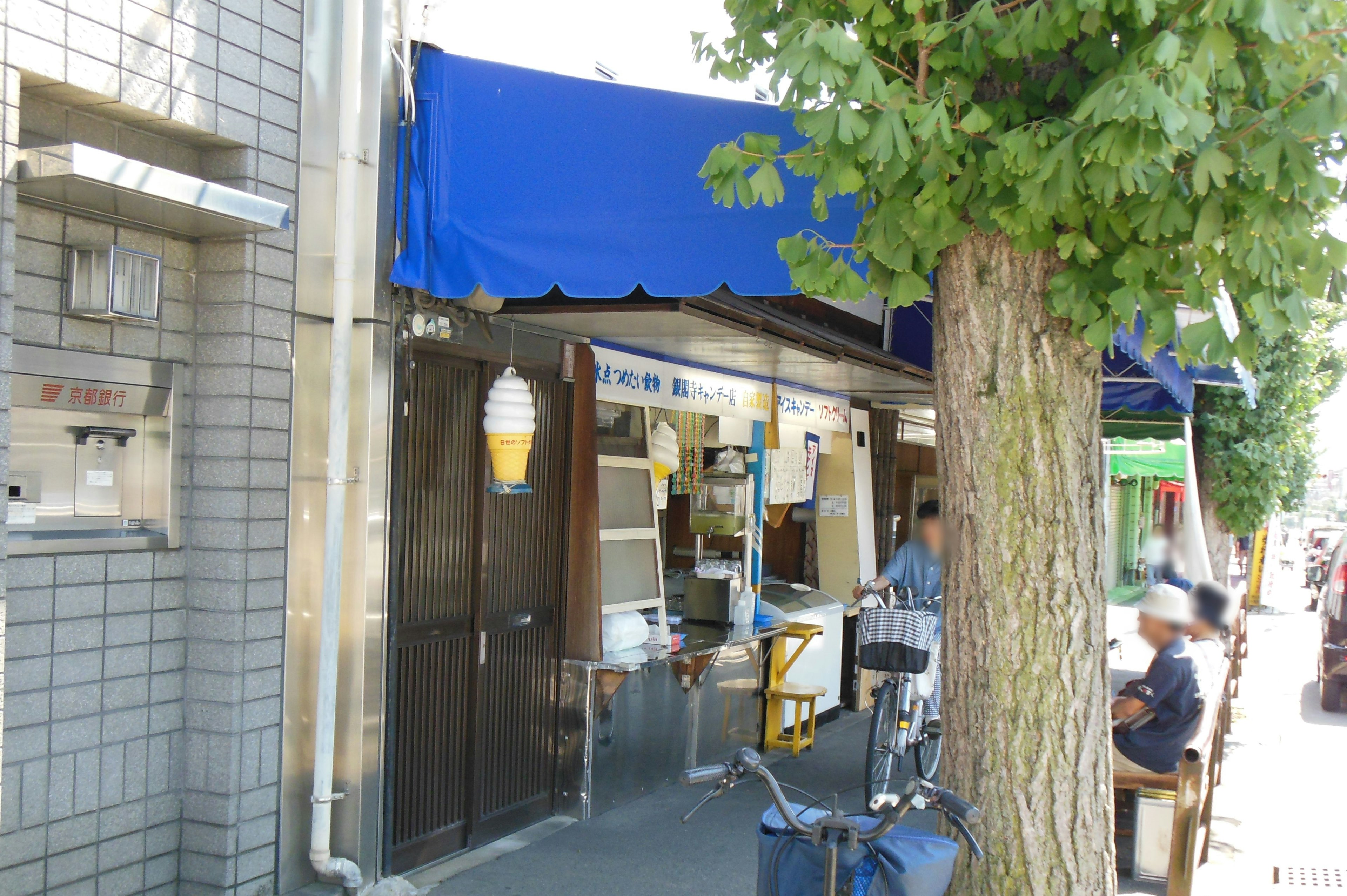 Exterior de un restaurante con un toldo azul rodeado de árboles y personas