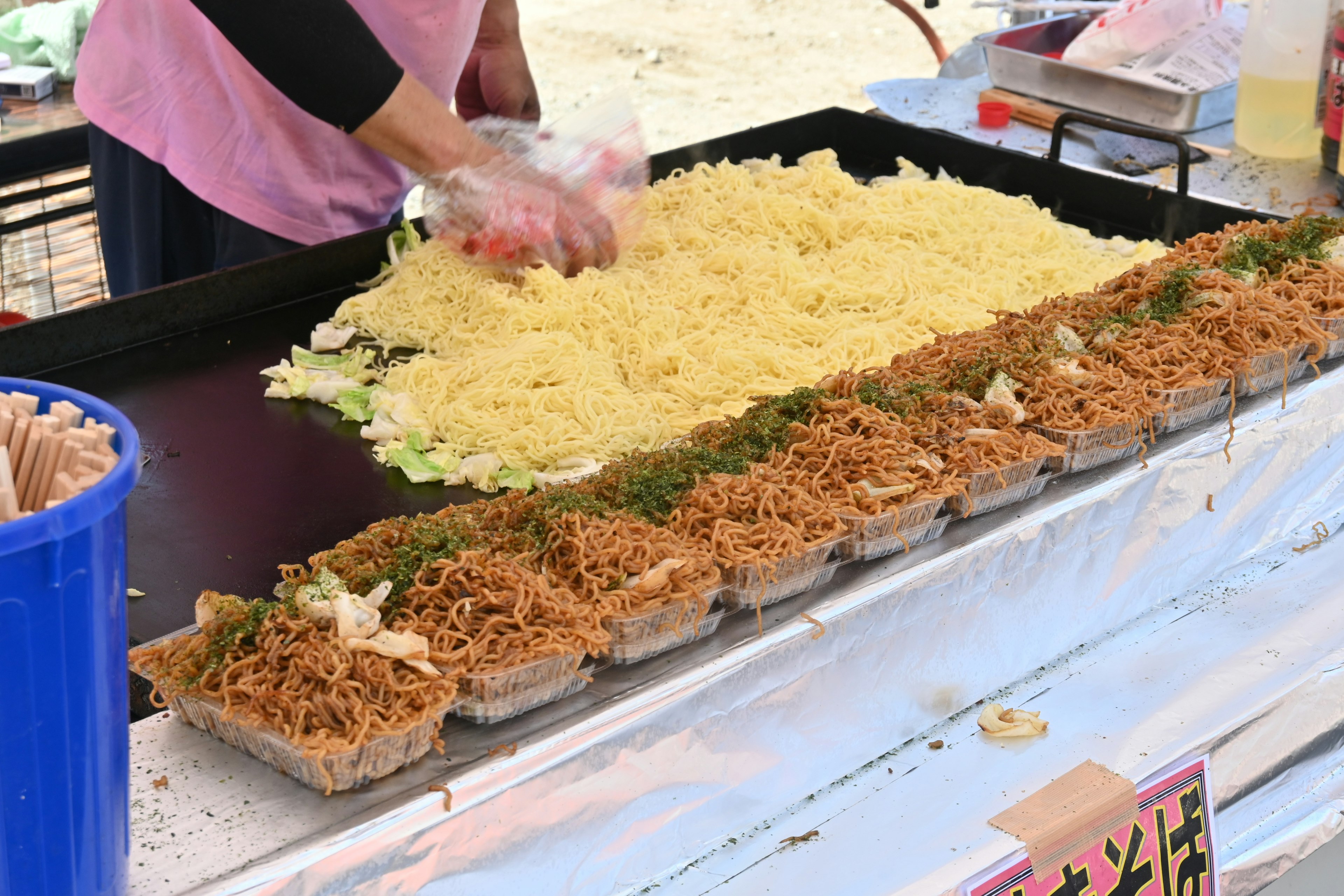 A vendor preparing yakisoba on a large griddle with noodles and vegetables