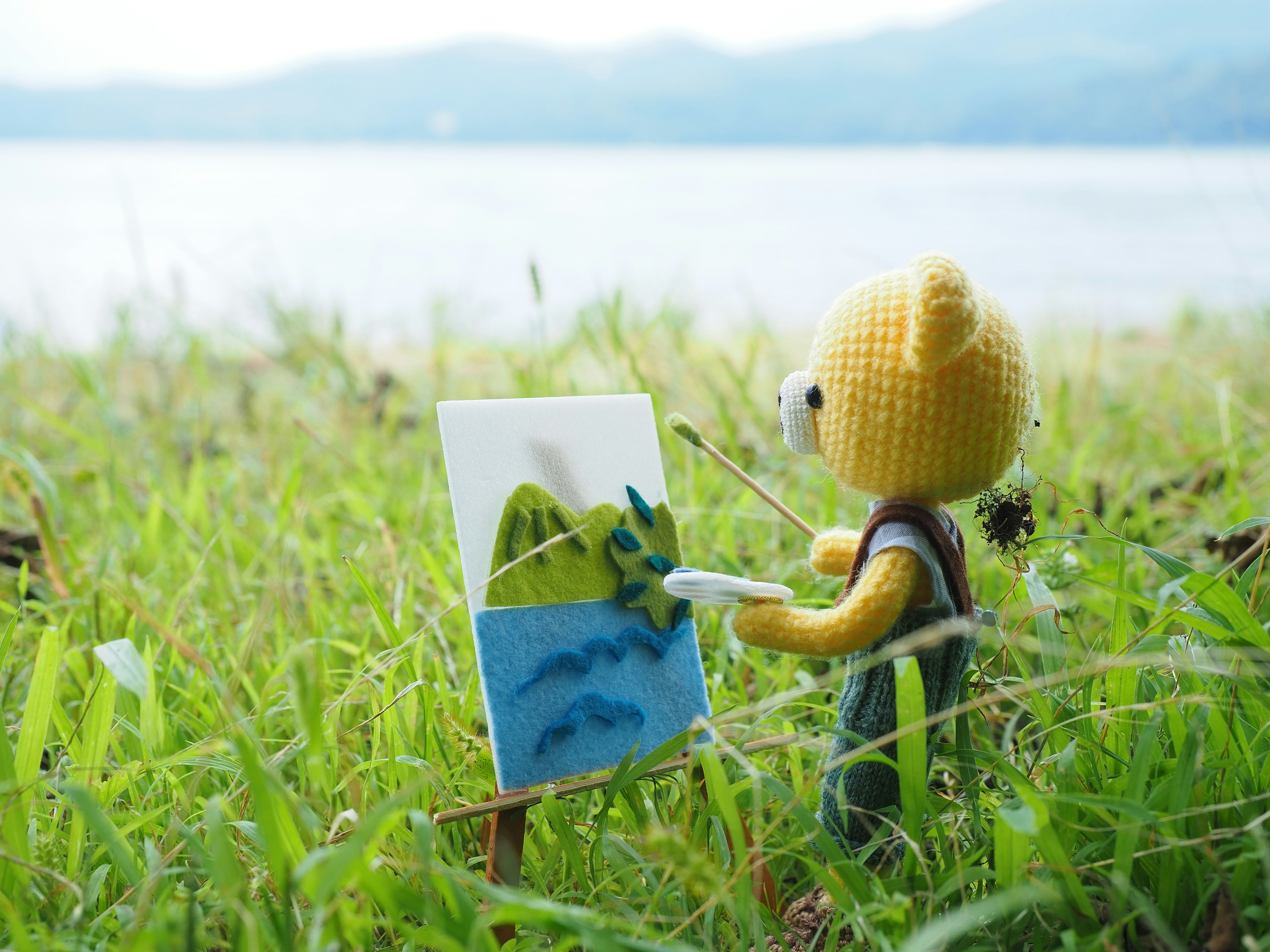 A plush bear character painting on a canvas in the grass by a lake