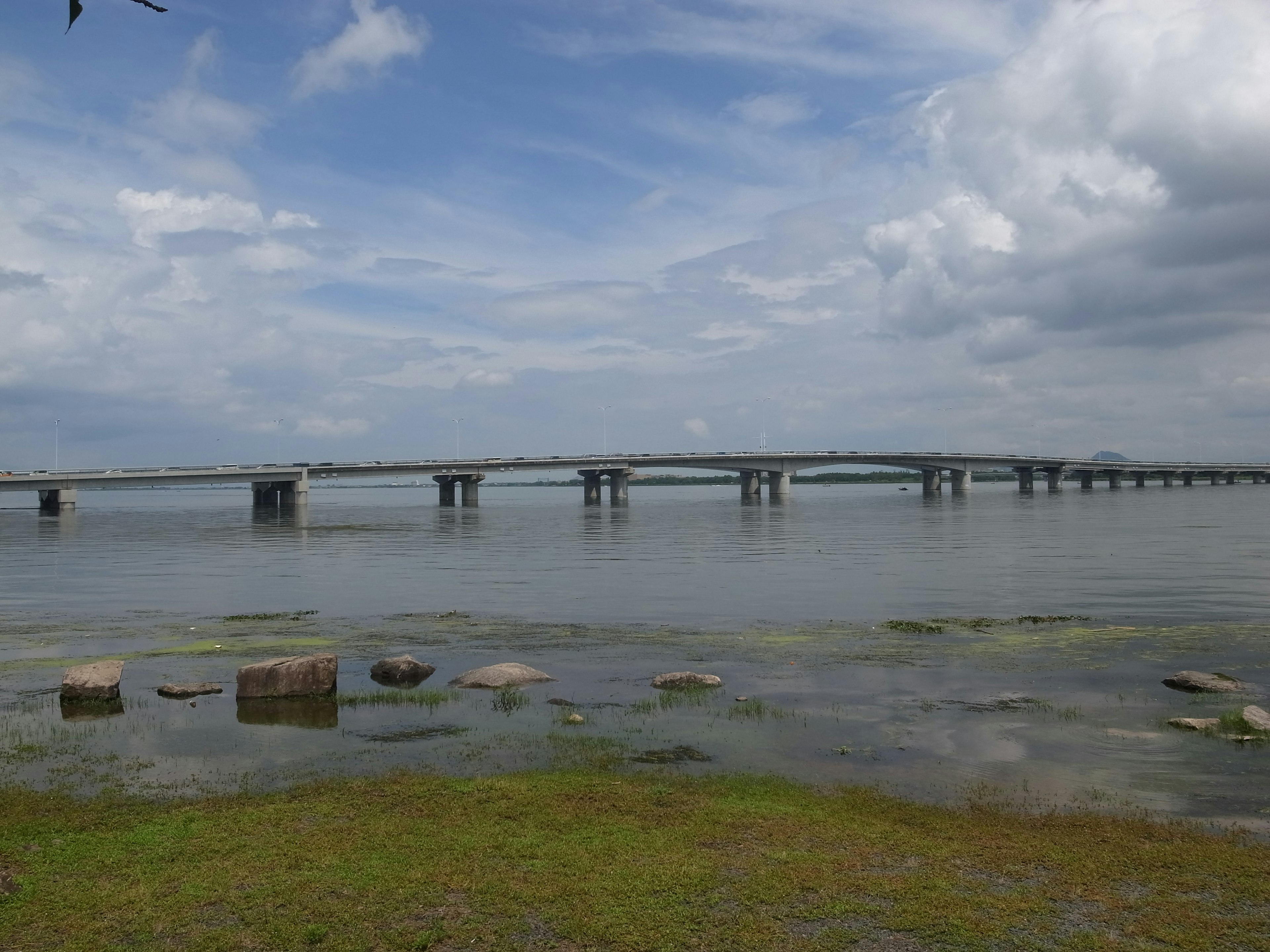 A bridge spanning over calm water under a blue sky with clouds and grassy shore