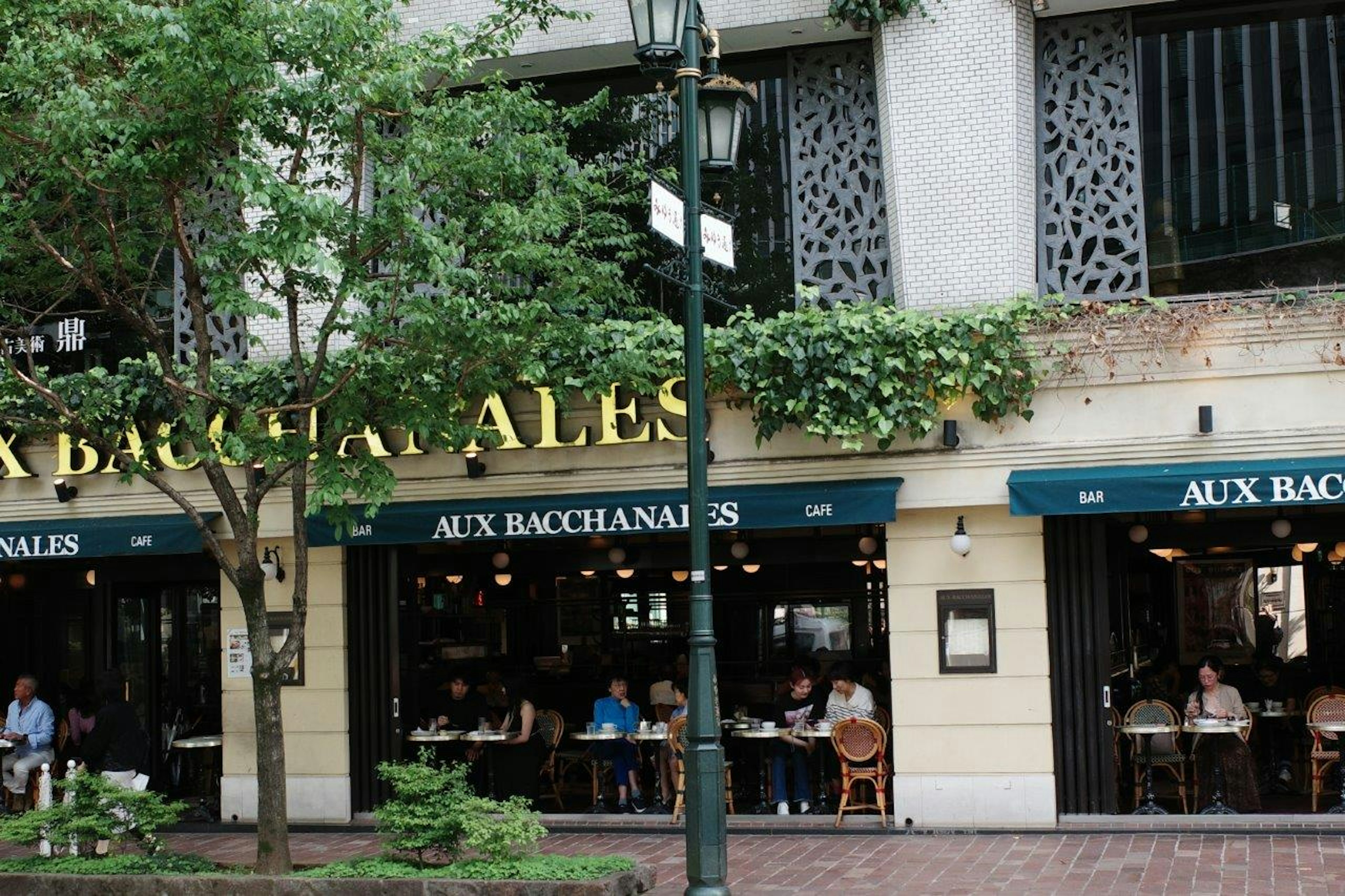 Vista exterior de un café con asientos en la terraza rodeados de vegetación que lleva el nombre "AUX BACCHANALES"