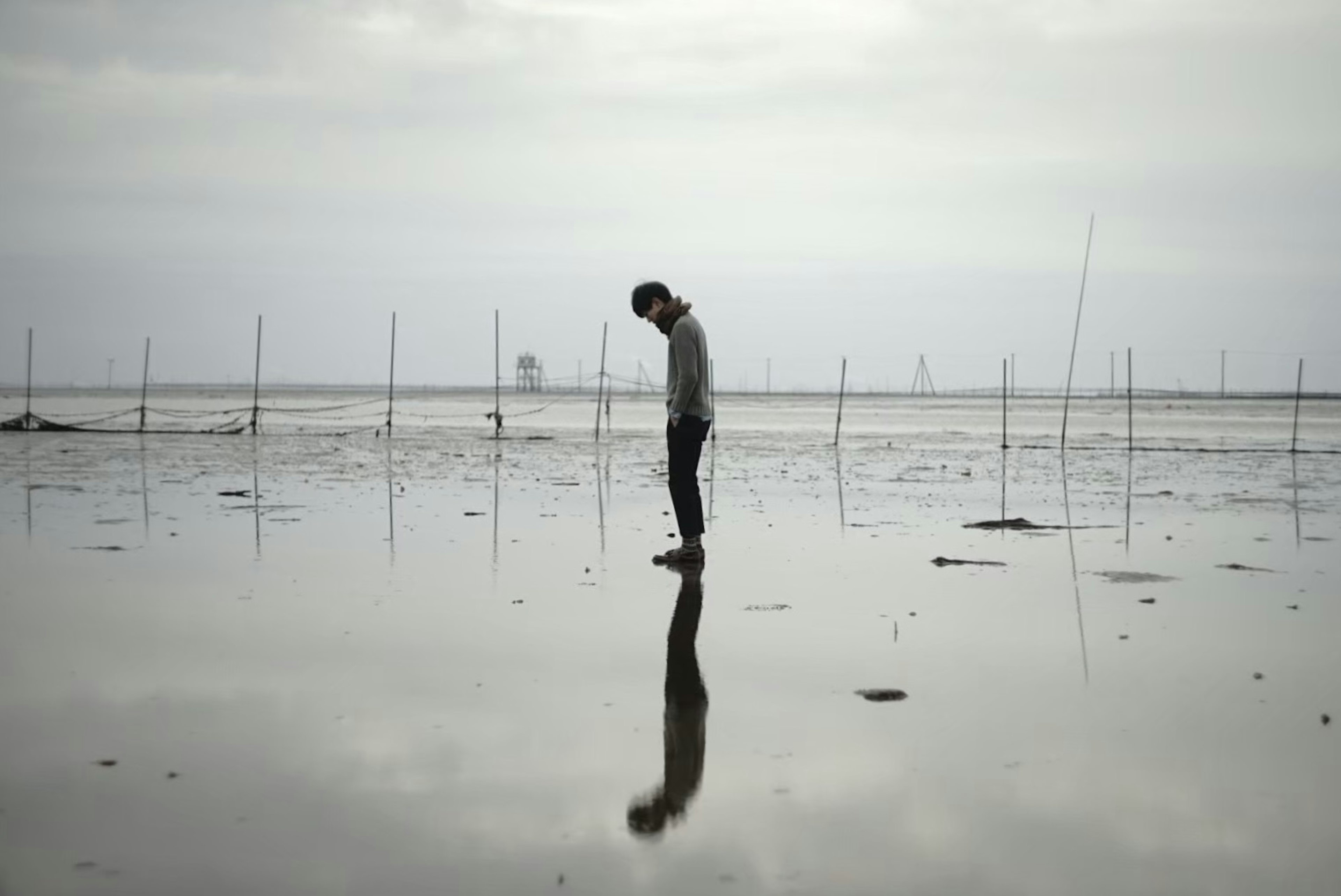 A figure standing alone on a reflective surface with poles in the background