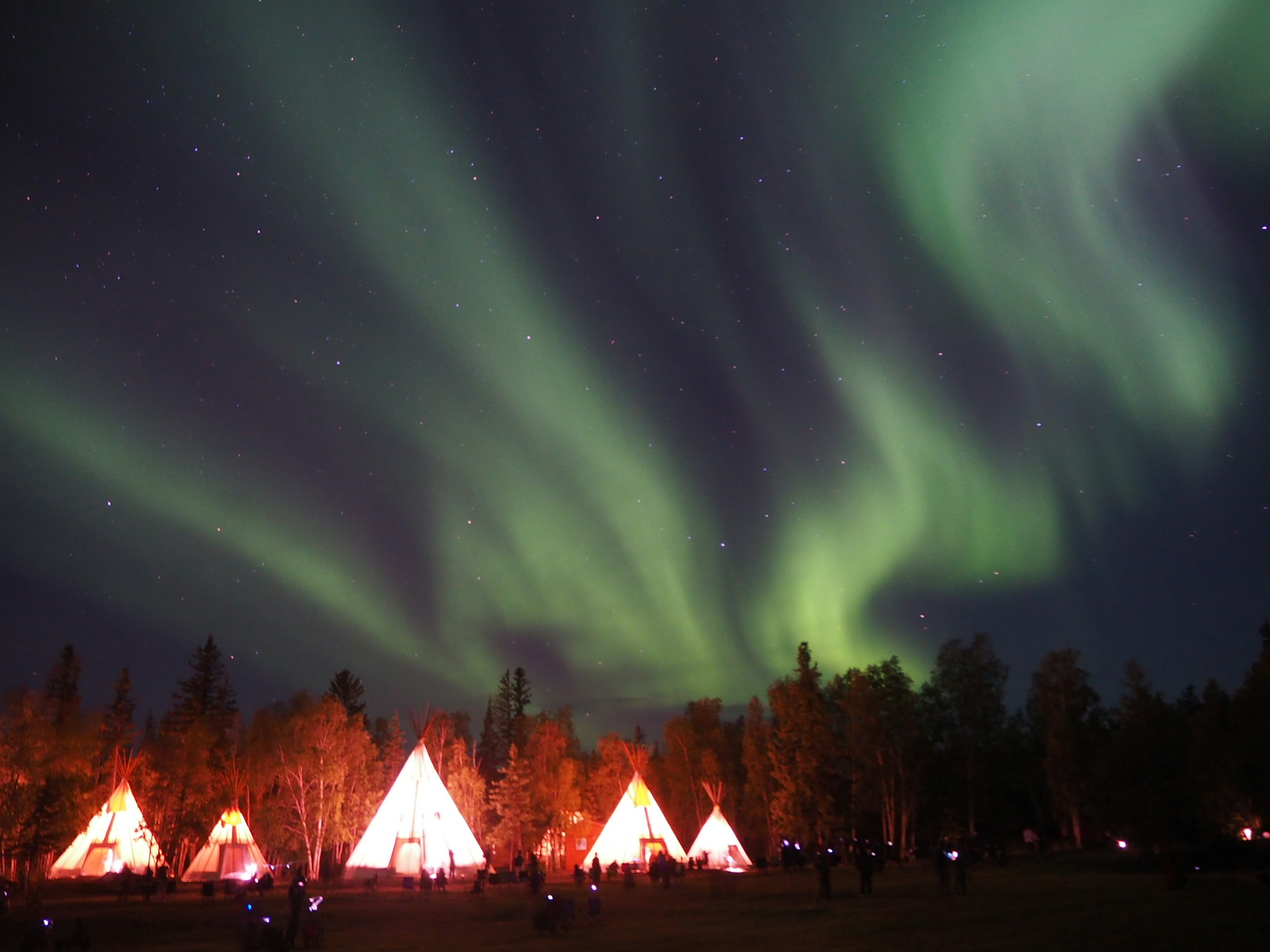 A beautiful aurora lights up the night sky above brightly lit tents