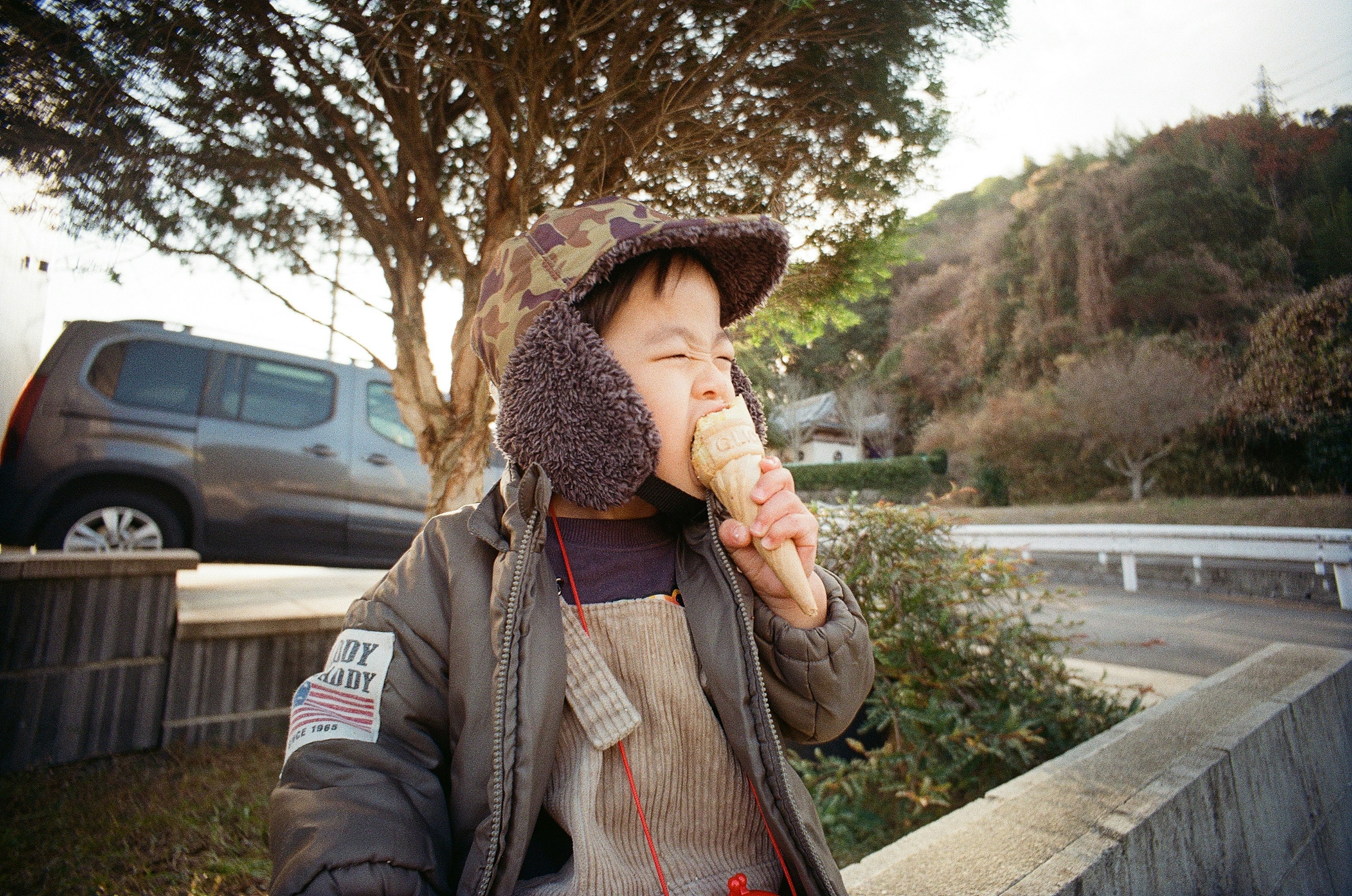 一個孩子在吃冰淇淋，戴著帽子和外套，背景有一棵樹和一輛車